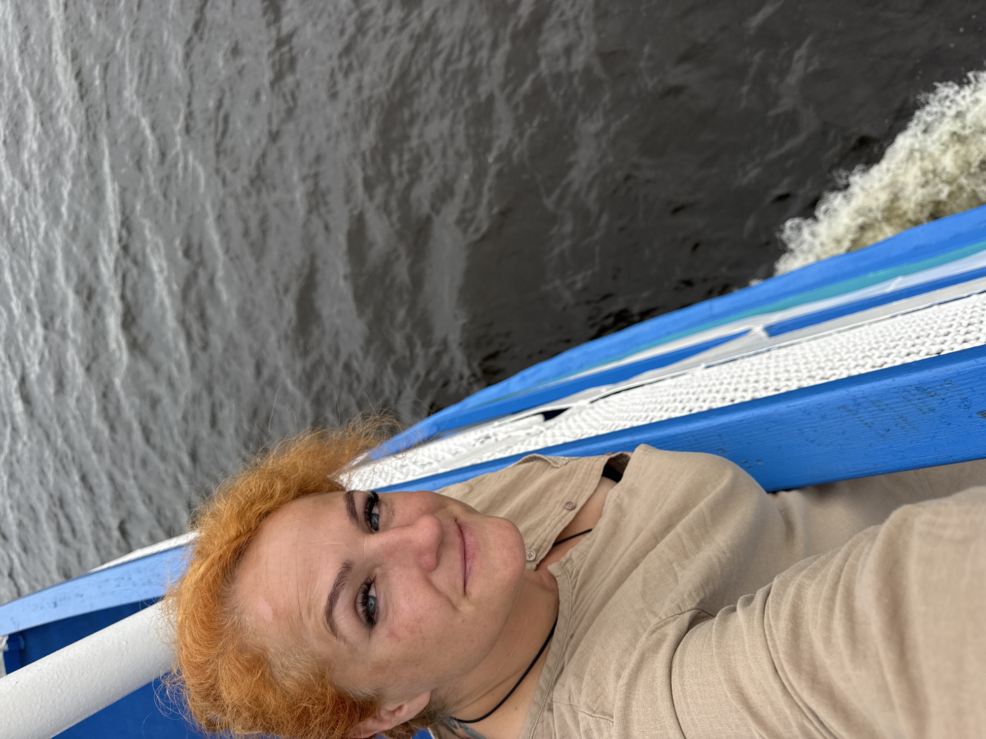 A woman with red, curly hair is taking a selfie on a boat. She is wearing a tan, button-up shirt and a black necklace. She has blue eyes, makeup, and a light tan complexion. The boat has a blue and white railing. The water is dark gray and choppy, indicating the boat is moving. The scene is likely outdoors, on a body of water.