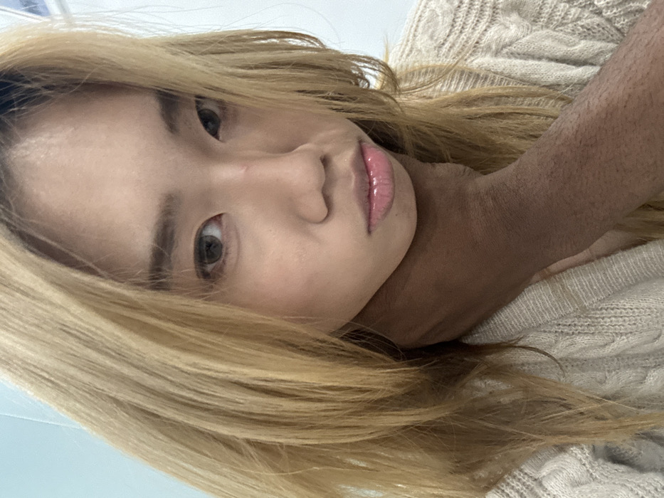 The image depicts a close-up portrait of an East Asian female, reportedly situated indoors in Yuzhong District, China. The female subject has light brown, shoulder-length hair and dark eyes. She is wearing a light-colored, cable-knit sweater. A faint, linear scar is visible on the bridge of her nose. Her lips are slightly parted and appear glossy. Her gaze is directed straight at the camera, and her facial expression is neutral.
Her chin and lower jaw are resting on a dark-skinned forearm, which enters the frame from the lower left. This forearm, exhibiting fine dark hair, and a portion of a dark-skinned hand are positioned against the female subject's throat and jawline, appearing to support or cradle her head. The remainder of the individual to whom the forearm belongs is not visible. The background is a plain, out-of-focus, light-colored wall, likely white or light blue. Lighting appears even and diffused.