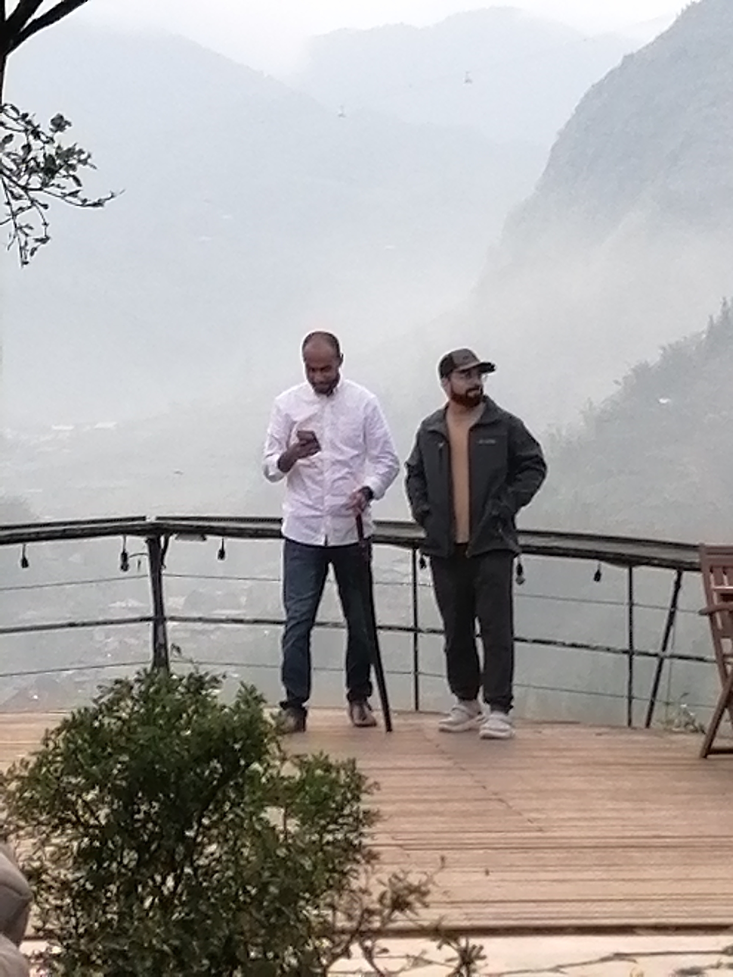 Two men stand on a wooden deck with a railing, against a backdrop of hazy mountains. The man on the left is fair-skinned, bald, wearing a white button-down shirt, blue jeans, and brown shoes. He is holding a smartphone in his hands and a cane in the other. The man on the right is fair-skinned with a beard, wearing a baseball cap, a grey jacket over a tan shirt, dark pants, and light-colored sneakers. He has his hands in his pockets. A green bush is in the foreground. The location is Sa Pa Ward, Vietnam.