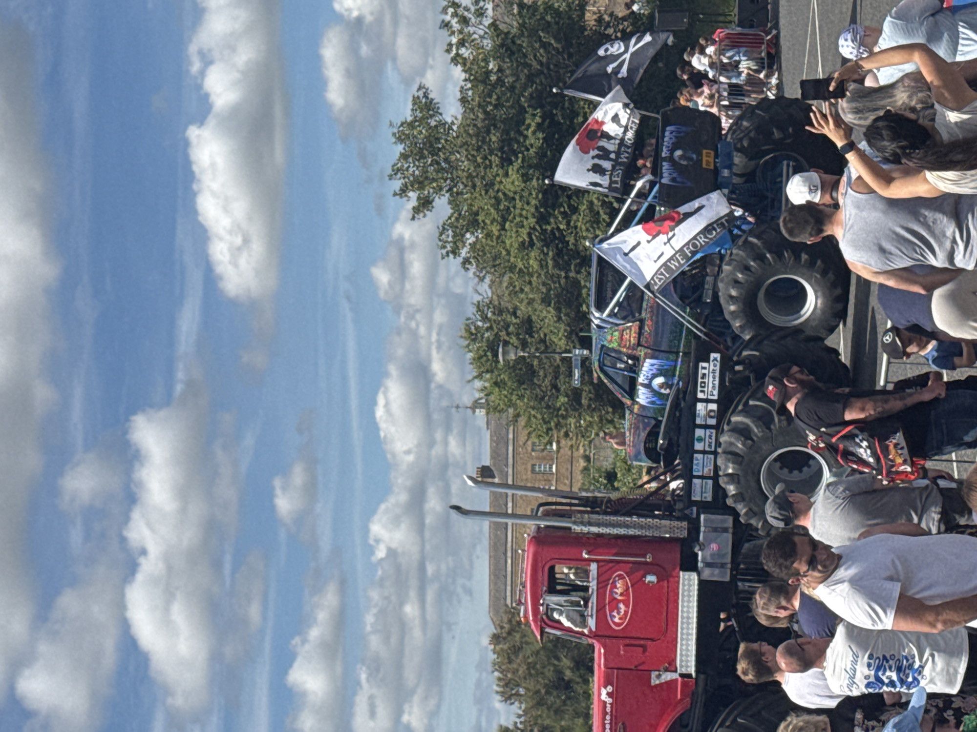 The image shows a large red monster truck and a second, darker monster truck with colorful graphics parked side-by-side on a paved area. Both trucks have enormous tires. The red truck has two people visible in the driver's cab, and a logo reading "Rip Roar" on its side. The darker truck has several flags attached to it, including one with a poppy and the words "LEST WE FORGET," and another with a skull and crossbones. There is a crowd of people standing in front of and around the trucks, some watching, some taking pictures. A building with a light-colored facade and a pitched roof is visible in the background behind the trucks, surrounded by trees. The sky is blue with scattered white clouds. The scene is likely outdoors, possibly at an event or festival. The image was taken in Great Yarmouth, United Kingdom.