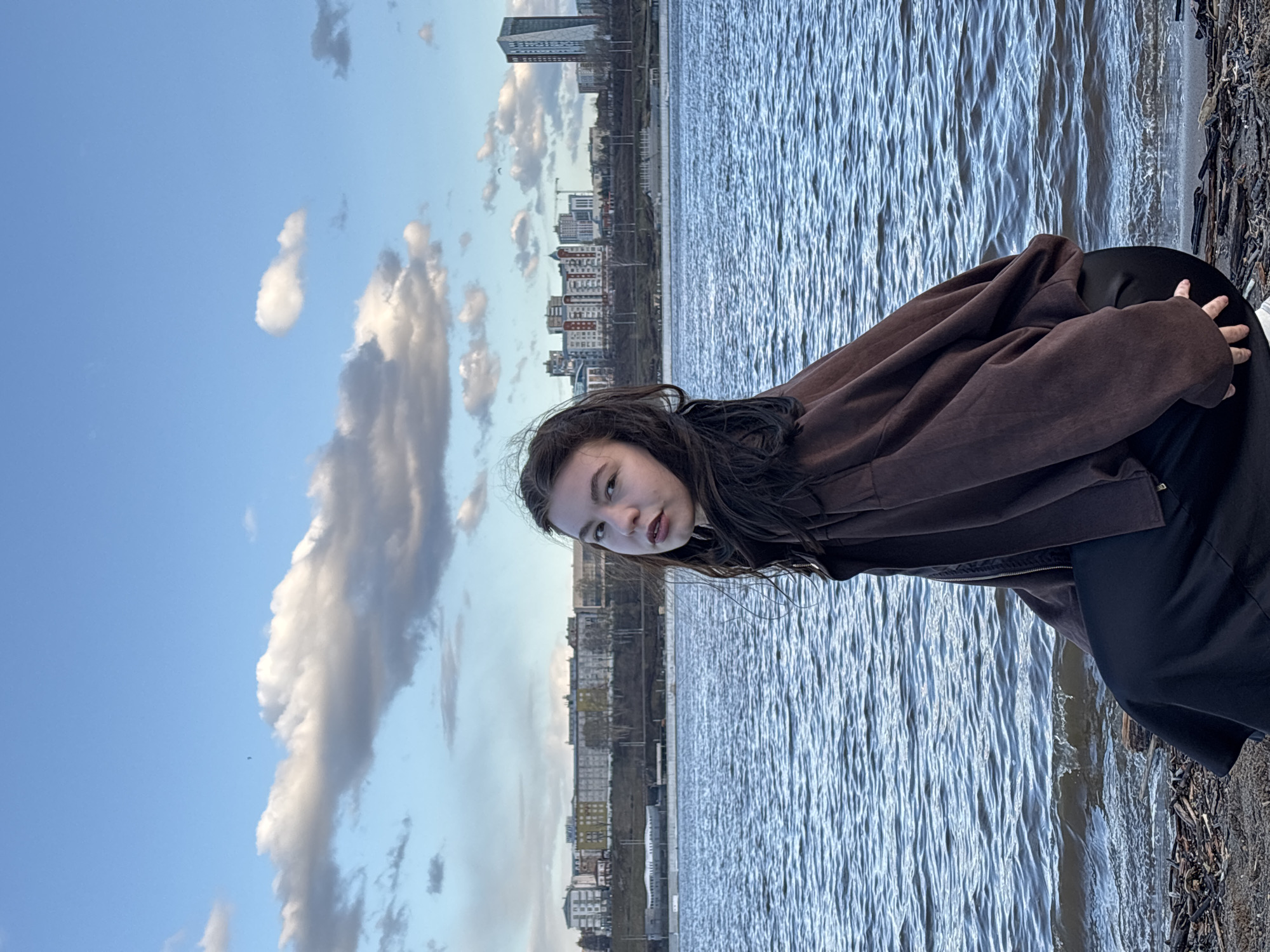 The image features a young female posing by a large body of water, likely a river, with a distant city skyline and a cloudy blue sky in the background. The photograph is taken from a slightly elevated perspective.
The main subject, a young woman with long, dark hair, occupies the left side of the frame. She is wearing a dark brown jacket over dark attire and is leaning forward, looking directly at the camera with a neutral expression. Her right hand rests on a dark, indistinct surface near the water's edge.
The middle ground is dominated by the expansive water body, which exhibits distinct ripples and reflections across its surface. Along the immediate bank to the right of the water, tangled dark debris, possibly driftwood or roots, is visible. Beyond the water, a cityscape stretches across the horizon, showcasing a mix of modern high-rise buildings and other structures.
Above the city and the subject, the sky is a vibrant blue, adorned with scattered clouds. These clouds range from small, bright white formations to larger, darker grey ones, suggesting partly cloudy weather. The lighting indicates it is daytime, possibly late afternoon, given the soft illumination. The scene conveys an urban natural setting, consistent with Perm, Russia, likely along the Kama River. No visible text is present in the image.