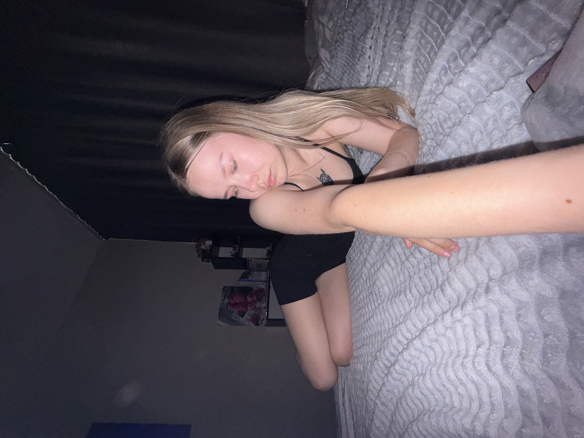 A young woman with blonde hair is lying on her side on a gray textured bedspread. She is wearing a black tank top and black shorts. Her eyes are closed, and she appears to be asleep or resting. She has a small tattoo on her chest. Her arm is extended towards the camera. A black curtain is visible in the background on the left side of the image. A small shelf with items is behind her. The environment appears to be a dimly lit bedroom. The location is unspecified and not apparent from the image; therefore, Chaikovsky, Russia cannot be confirmed.