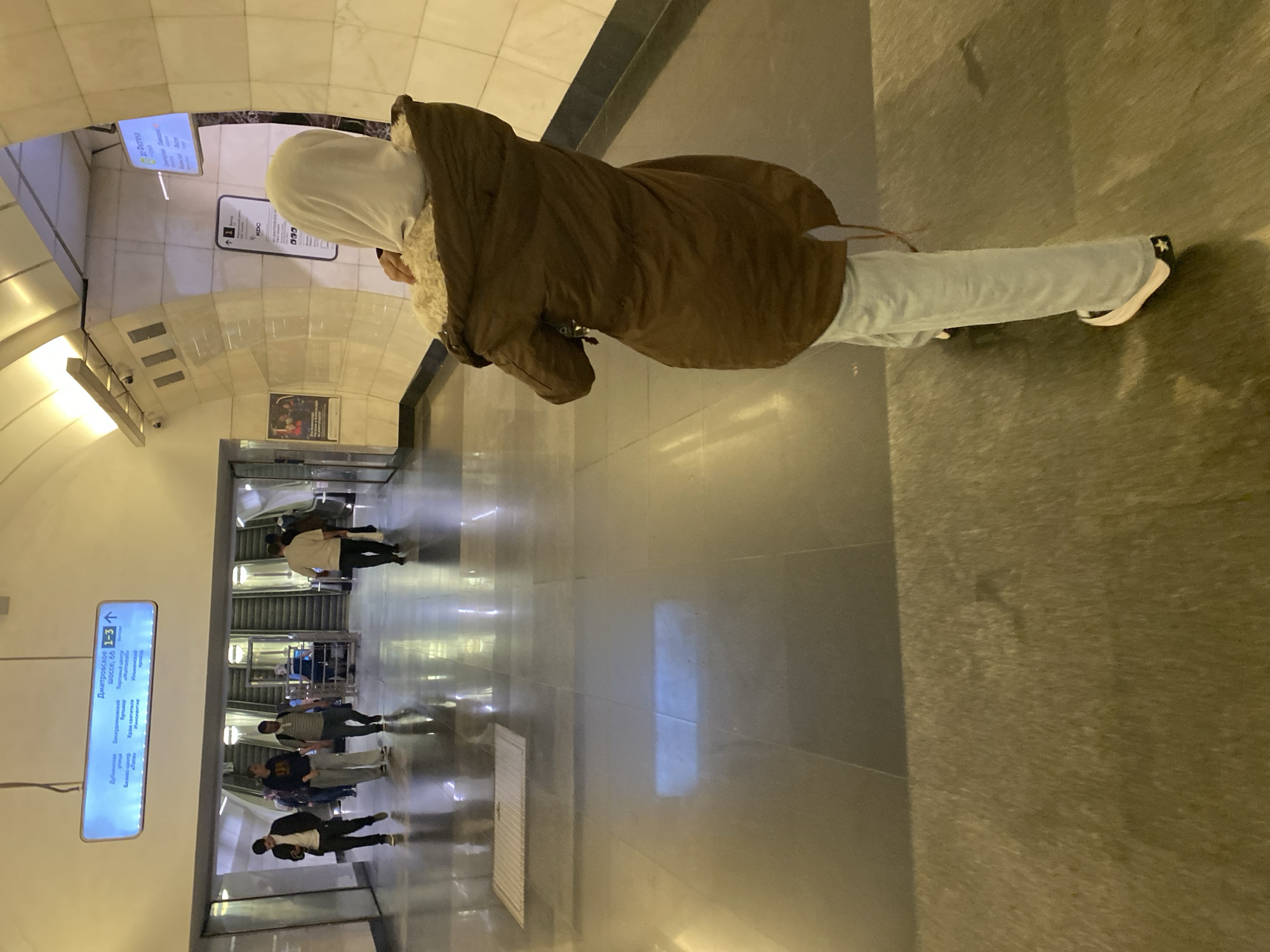 This media file depicts a person, viewed from the back, standing in a modern subway station in Moscow, Russia. The person is wearing a brown hooded coat with a faux fur lining and light-colored pants. They are facing a wall with a digital display that shows directions and station names in Russian. Other people are visible in the background, walking on the polished floor or on escalators. The station appears to be clean and well-lit, with reflective surfaces that create a sense of spaciousness. The overall atmosphere seems to be one of transit and daily life in a bustling urban environment. The visible text on the display includes station names like "Дмитровская" and "Дубровка," and directional arrows. The time of day is not explicitly indicated, but the artificial lighting suggests it could be any time. There are no weather cues visible.