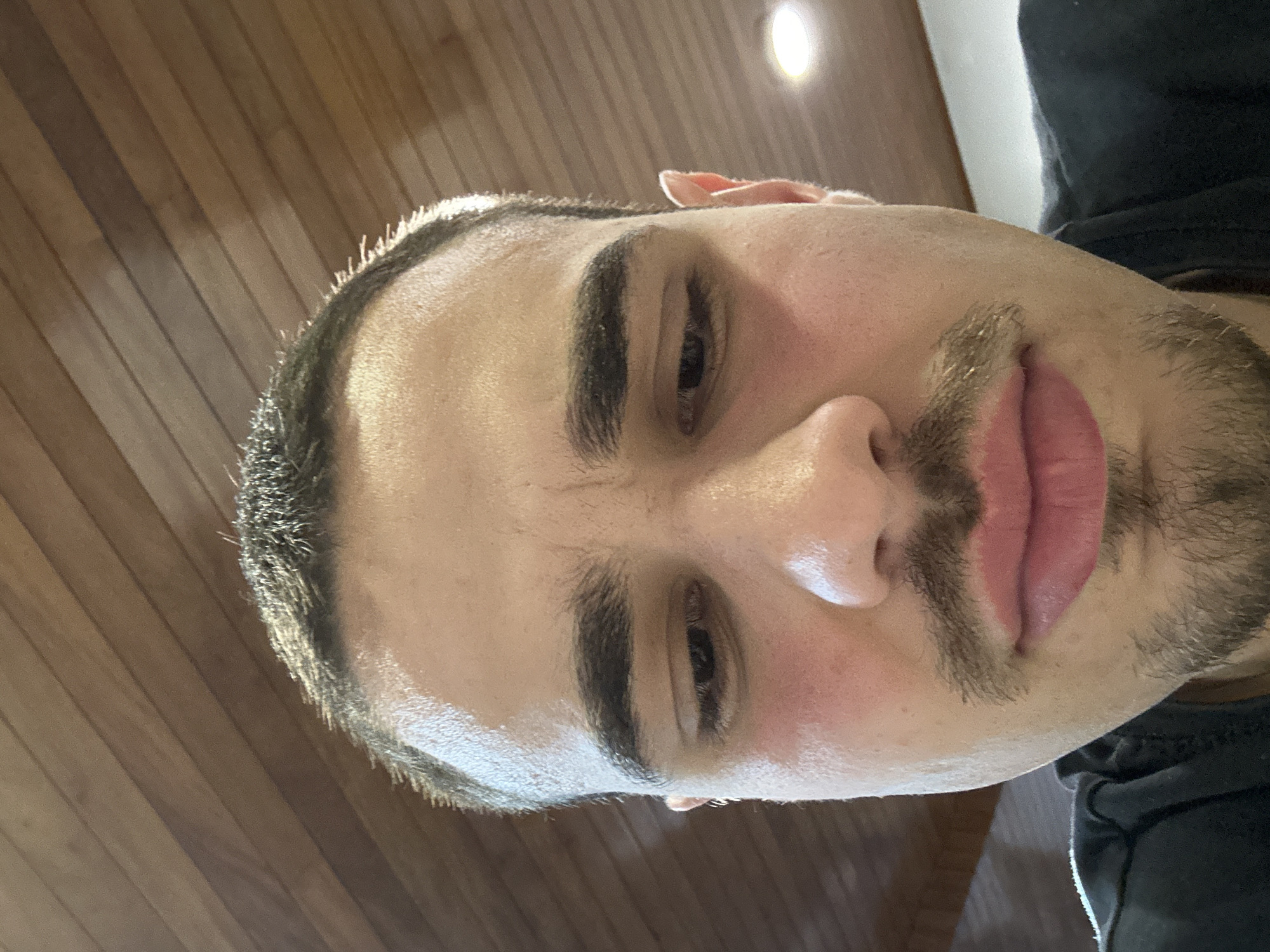 A close-up, tilted, indoor photograph depicts a man's face. He has dark hair, short and close-cropped, with dark eyebrows and stubble. The man's skin appears light and slightly flushed. The background consists of a light-colored wooden surface. The man is wearing a dark-colored shirt, visible in the lower right corner. There is no information to place the image location in Mirandela, Portugal.