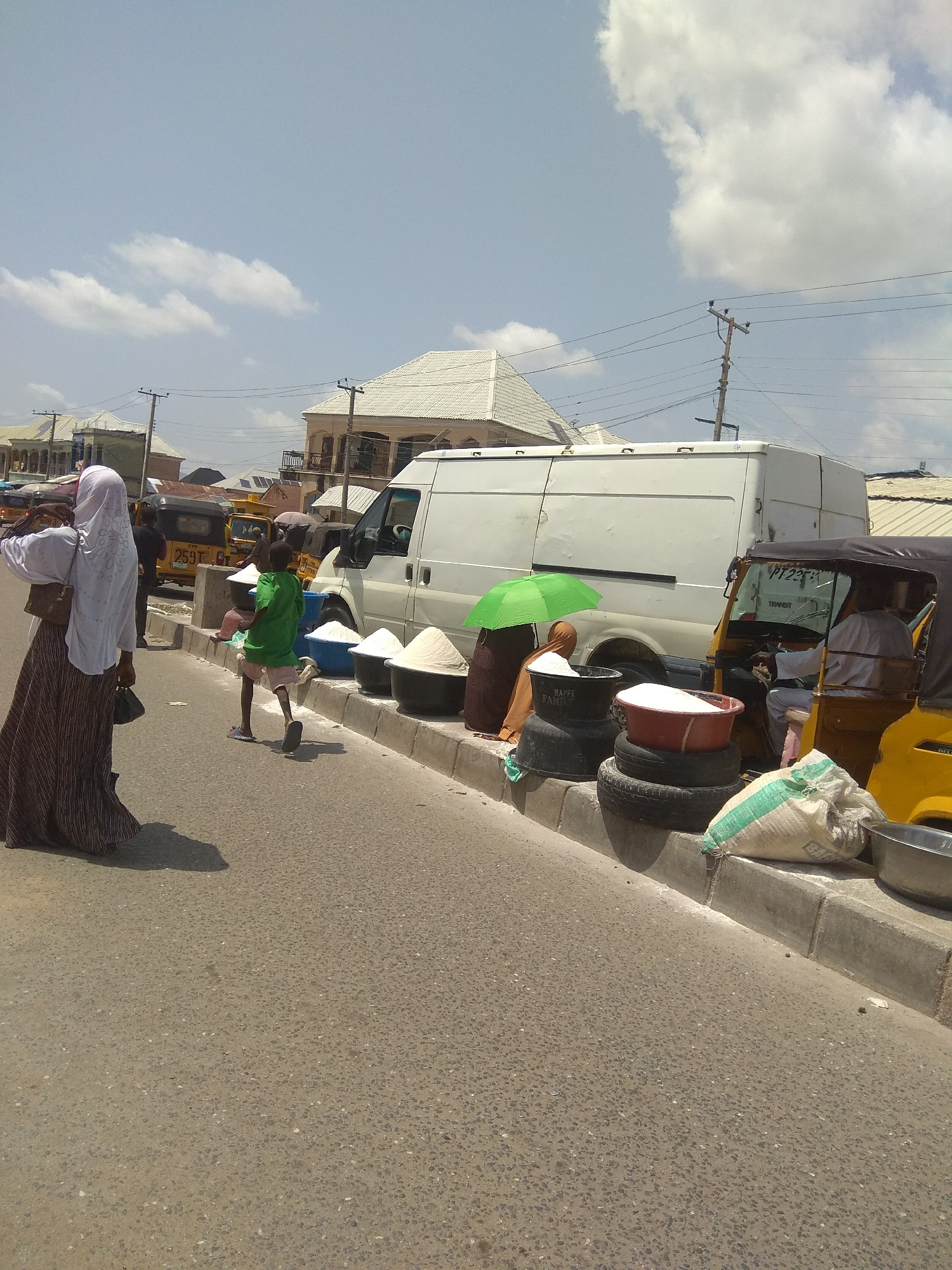 The image depicts a bustling street scene in Jalingo, Nigeria, under a bright, partly cloudy sky, suggesting it is daytime. The foreground is dominated by a paved road with a concrete curb running along its edge. Several large, black tires are stacked on the curb, serving as a makeshift base for large, dark bowls filled with a white granular substance, likely rice or sugar, being sold.
To the left, a woman wearing a white hijab and a long, patterned skirt walks away from the camera, carrying a dark bag and a smaller dark object in her hands. Beside her, a young boy in a green t-shirt and light-colored shorts runs across the road, looking towards the right.
In the middle ground, a large white van is parked alongside the curb, possibly a delivery vehicle. Beside it, another vendor, a person in a brown outfit, is seated under a green umbrella, attending to their goods. A yellow three-wheeled vehicle, a common mode of transport in Nigeria, is also visible, with a person seated in the driver's seat. Several other similar vehicles are parked further down the street, indicating a busy marketplace or transport hub.
In the background, buildings with white roofs and balconies line the street, typical of the region. Electrical poles and wires crisscross the sky. One of the bags of produce near the yellow vehicle has green and white stripes. The overall atmosphere appears active and vibrant, characteristic of a local market day. No specific emotions are overtly conveyed by the individuals, but the scene suggests daily commerce and movement.