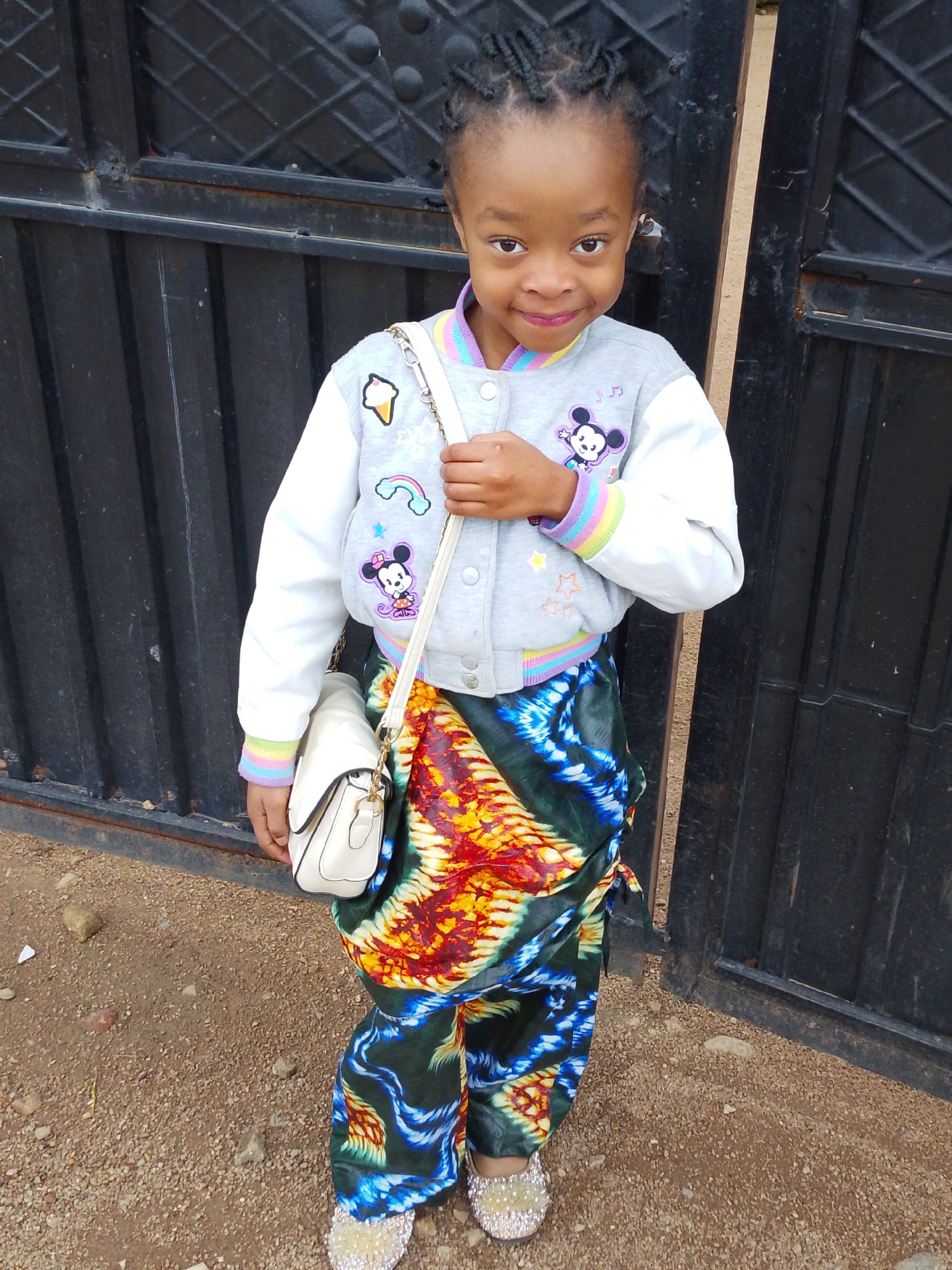 A young girl stands in front of a black metal gate. She is wearing a light gray jacket with white sleeves, adorned with various patches, including a Mickey Mouse motif and a rainbow. Her lower body is covered in a vibrant, patterned fabric that wraps around her like a skirt or sarong, featuring bold colors of yellow, orange, blue, and black. She carries a small, white purse with a gold chain strap over her shoulder. The ground is covered in small gravel and dirt. The background consists of the textured black metal gate. The lighting suggests it is daytime. The scene is likely outdoors, possibly in a residential area within Jos, Nigeria, given the typical architecture and environment found in the city.