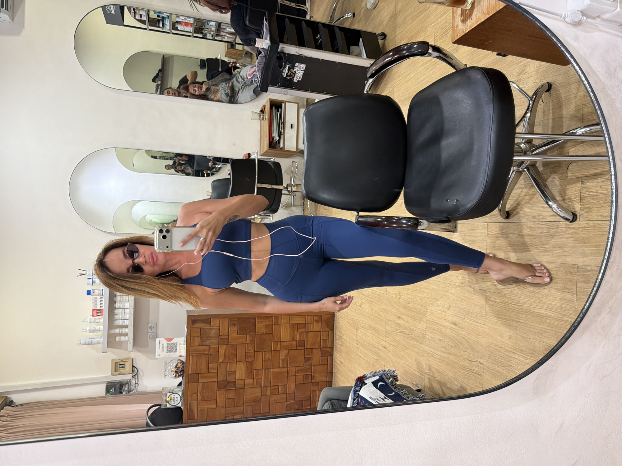 A woman wearing sunglasses and a navy blue workout set is taking a selfie in a mirror within a salon in North Kuta, Indonesia. She has blonde hair and is holding a smartphone in her right hand. In the background, reflected in the mirror, are salon chairs, mirrors, shelves with various products, and other people receiving hair services. The salon appears to have wooden floors and walls.