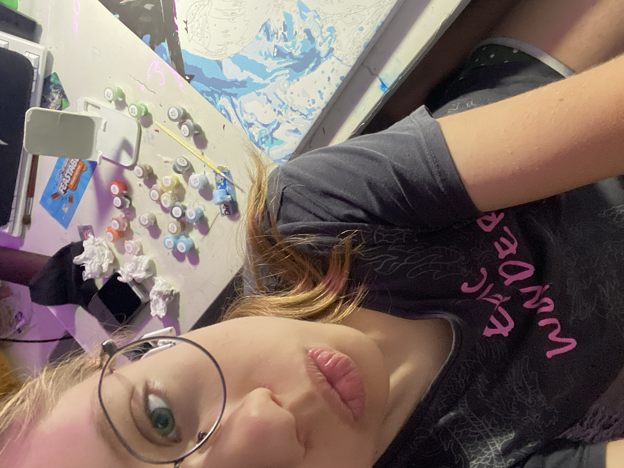 A close-up, high-angle selfie shows a young person with blonde hair, wearing glasses and a dark t-shirt with pink lettering that reads "epic wonder." They are in a room, with a desk covered in supplies for a paint-by-numbers kit. Small containers of paint, brushes, and crumpled paper are scattered across the desk. A large canvas with a partially completed painting in shades of blue and white is visible to the right. The background is dimly lit with purple and pink lighting. The person's left arm is resting on the desk, and their legs are visible in the lower right corner, wearing dark shorts with a green polka dot pattern. The image does not contain any information that would allow for the determination of its location within Zassovskaya, Russia.