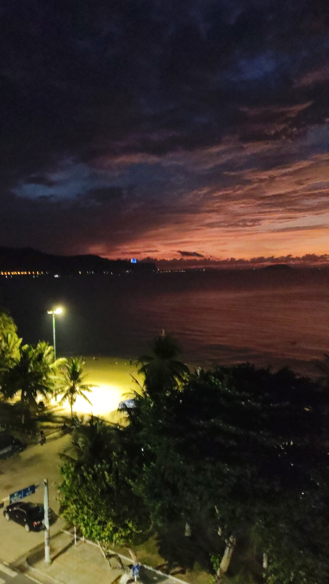 The image shows a nighttime scene in Bac Nha Trang Ward, Vietnam. A coastline stretches across the middle ground, with water illuminated by the sunset. The sky above is filled with dark clouds, but a reddish-orange hue persists at the horizon. In the foreground, trees line a street alongside a sidewalk with at least one car visible. A street lamp illuminates palm trees. Lights are visible along the coastline in the distance.