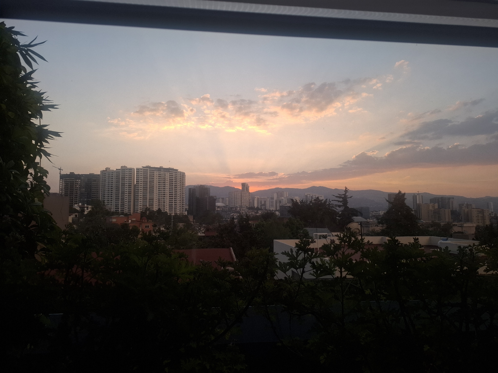 The image shows a view from a window in Huixquilucan de Degollado, Mexico, overlooking a cityscape at sunset. In the foreground, foliage obscures the lower portion of the frame. Behind the trees, several buildings of varying heights are visible, including large apartment complexes and smaller residential structures. The sky is a mix of blue and orange hues, with sunlight breaking through clouds. Distant mountains are silhouetted on the horizon. A construction crane can be seen behind one of the buildings. The overall scene is illuminated by the fading light of the setting sun.