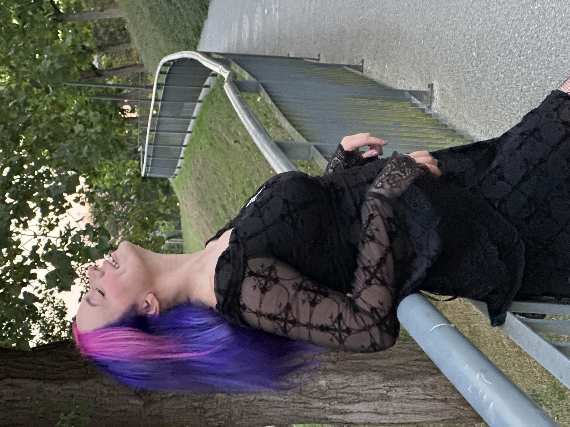 In Oviedo, Spain, a young woman with vibrant purple and pink hair leans against a metal railing, her head tilted back and eyes closed in apparent bliss. She wears a black lace dress with bell sleeves and is positioned near a tree on the left side of the frame. The background shows a paved pathway curving to the right, bordered by a grassy slope and a metal fence. The scene appears to be outdoors in a park or public area.
