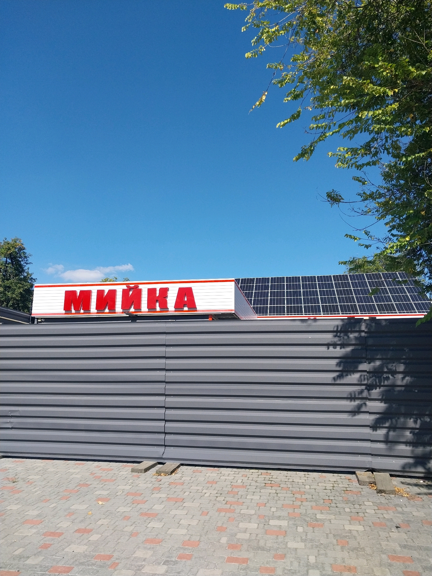 A close-up, eye-level shot of a car wash sign is captured under a clear blue sky. The sign, in bold red lettering, spells out "МИЙКА," which translates to "car wash." The sign is mounted on a white structure with a red trim, which is itself positioned above a dark gray, corrugated metal fence. Behind the sign and to the right, a section of a roof is visible, covered with numerous black solar panels. Green tree branches with visible leaves extend from the top right corner of the frame, casting shadows on the fence below. The ground in front of the fence is paved with interlocking rectangular bricks of varying gray and red hues. The scene appears to be in an outdoor, urban setting.