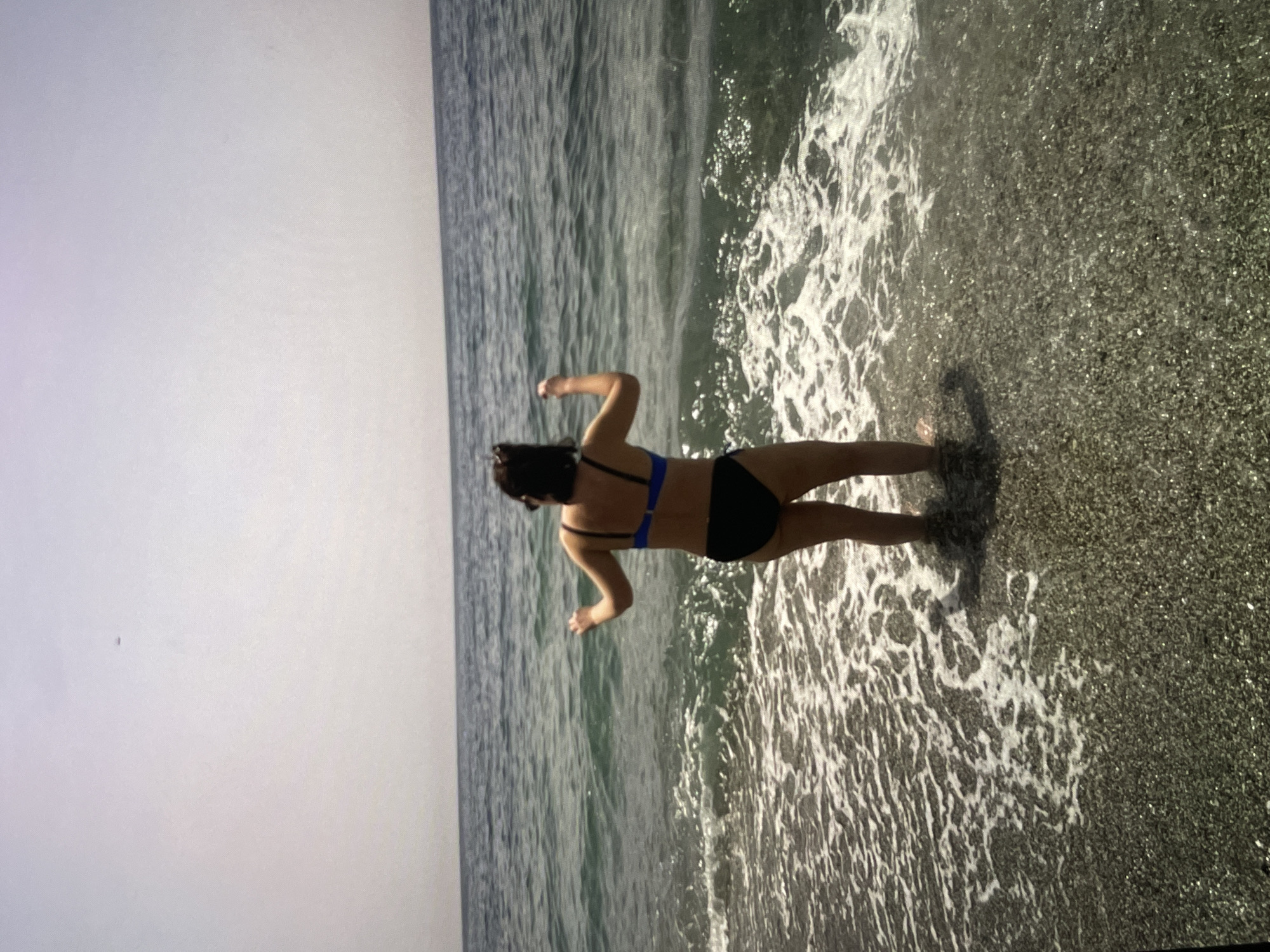 A woman in a blue and black bikini is standing at the edge of the beach in Unknown, Unknown. She is facing the ocean with her arms outstretched. The sky is overcast. The beach appears to be made of dark sand or pebbles. Waves are washing up to the shore.