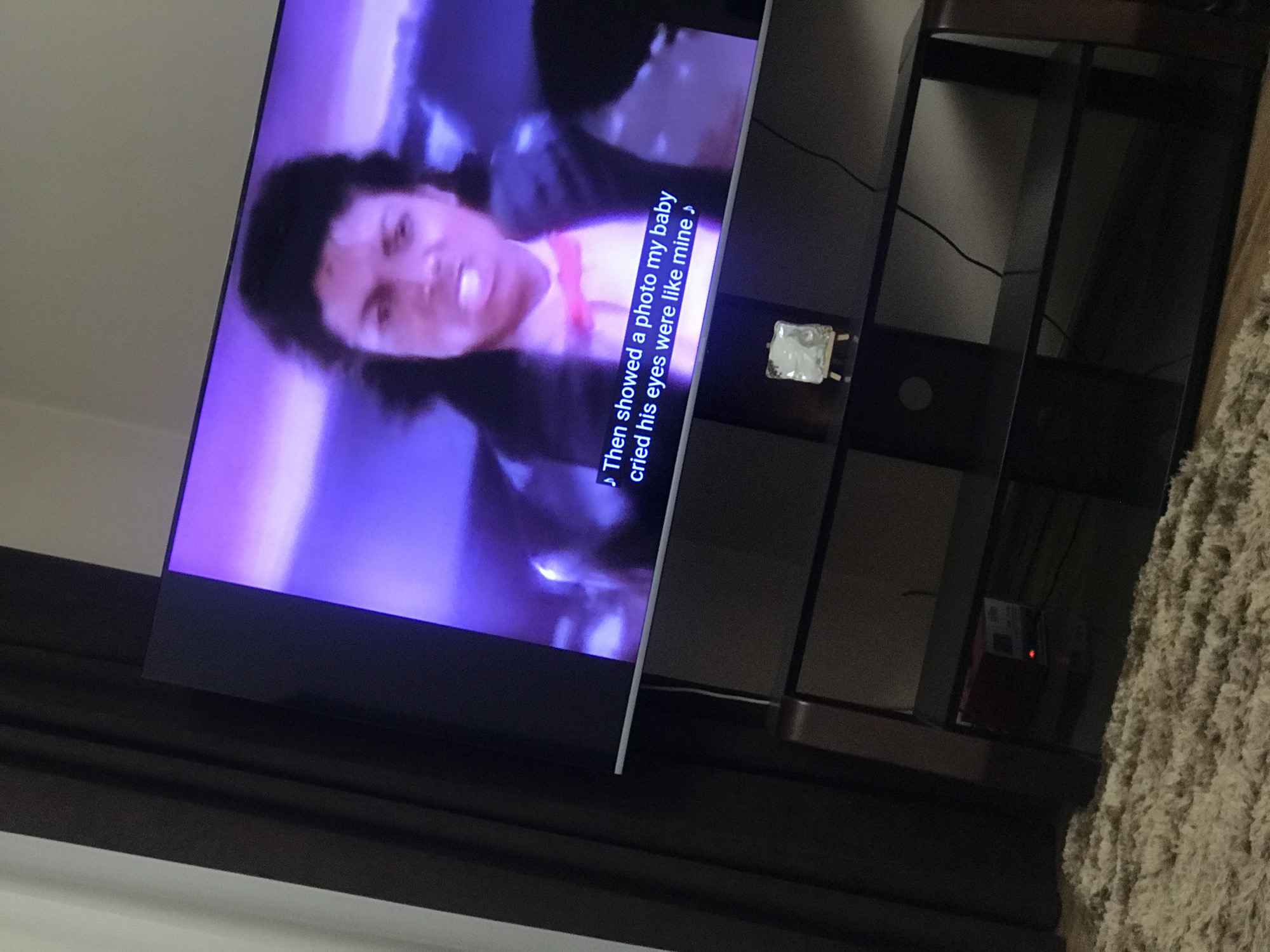 The image shows a television displaying a scene with a person and subtitles that read, "Then showed a photo my baby cried his eyes were like mine." The television is on a dark brown entertainment center. Adjacent to the entertainment center, there is a textured rug. The environment includes a wall and curtain. The location is not discernible from this image.