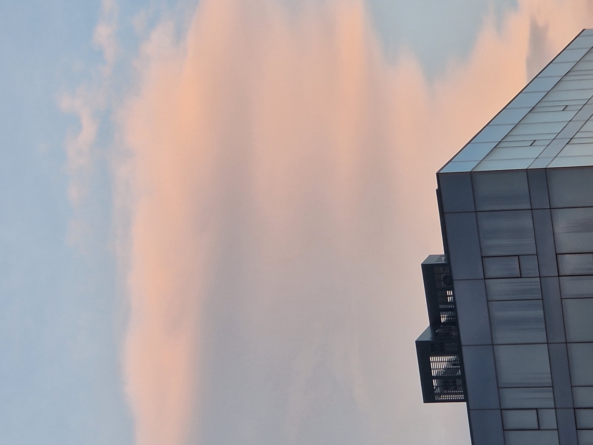 The image captures a modern building against a colorful sky, presented with a dynamic diagonal composition. The building occupies the right side of the frame, its structure rising sharply from the bottom-right corner towards the top-right. It features a distinctive, angular design with a sloped upper section made of reflective panels, suggesting glass or polished metal. The main body of the building is clad in dark, rectangular panels arranged in a grid pattern. Two prominent, dark, grated structures protrude from the left side of the building, possibly utility enclosures or balconies.
The sky dominates the left and central parts of the image, showcasing a striking gradient of colors. On the far left, the sky is a soft, light blue, with faint, wispy clouds. Towards the center and right, the sky transitions into warm, delicate hues of peachy-pink and orange, indicative of either sunrise or sunset. A large, soft cloud mass reflects these warm colors prominently.
No people or discernible activity are present in the scene. The image has a calm and serene atmosphere, emphasizing the architectural lines of the building against the natural beauty of the sky. There is no visible text.