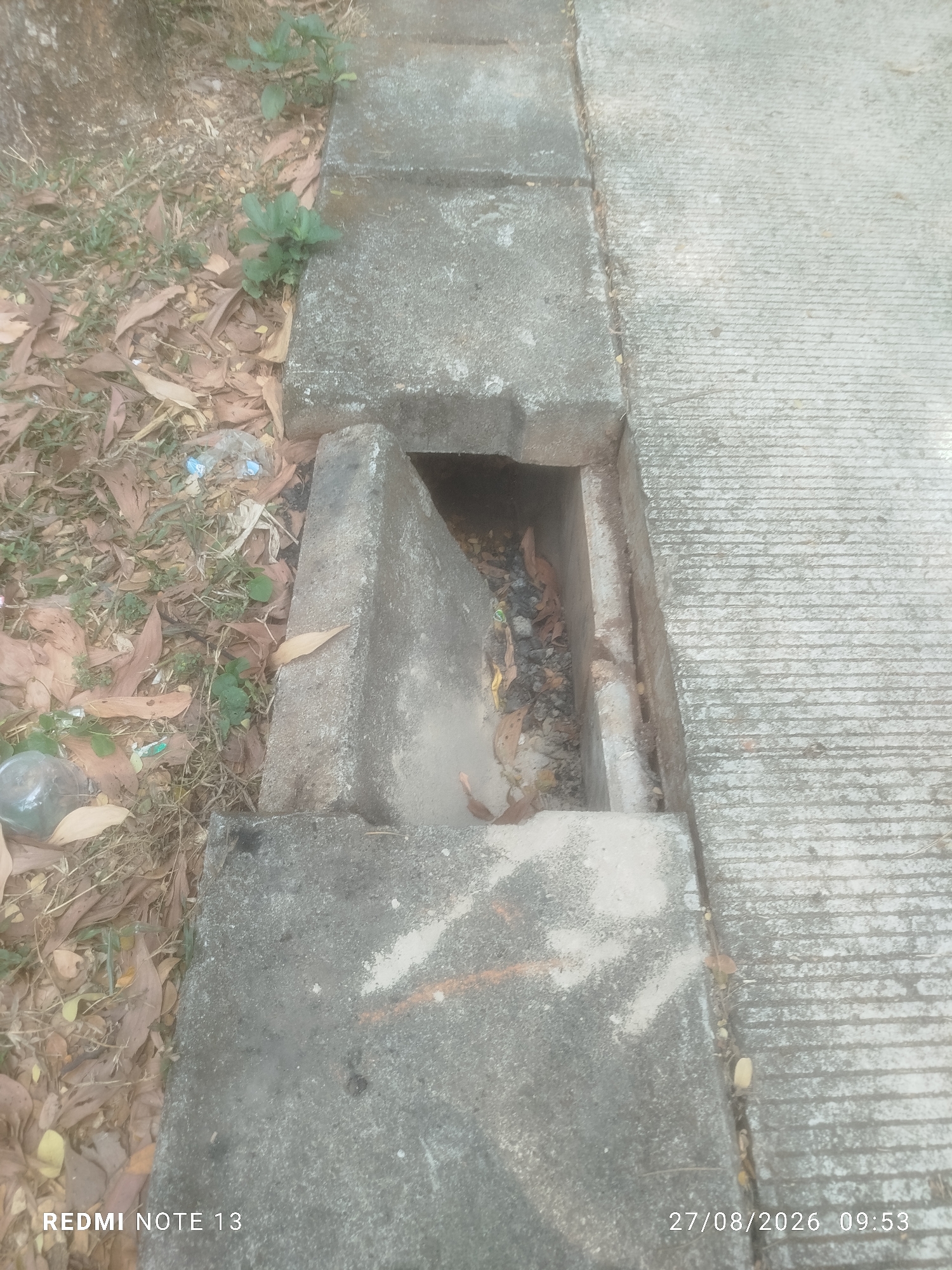 The image shows a concrete storm drain with debris inside, located on a sidewalk in South Tangerang, Indonesia. Leaves and some plastic waste are visible within the drain. The surrounding area consists of a concrete sidewalk and a grassy area with scattered leaves. The timestamp "27/08/2026 09:53" and "REDMI NOTE 13" are overlaid on the image.