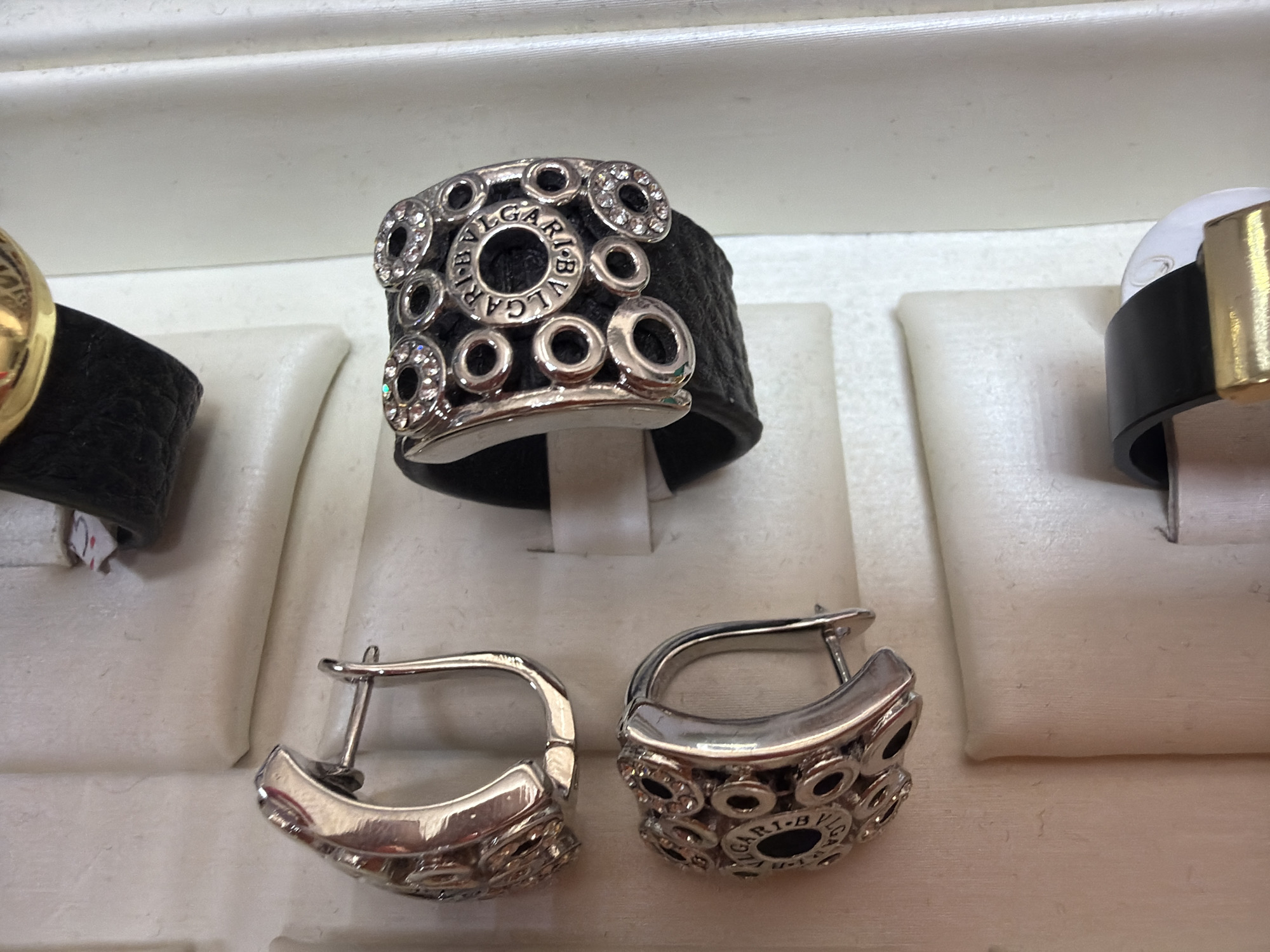 The image displays a close-up view of several pieces of jewelry, likely within a display case. The jewelry includes two silver earrings with intricate designs and a ring with a black band adorned with multiple circular metal accents and small clear stones. The background is a muted, textured surface, possibly fabric or padding, designed to showcase the items. The overall lighting is bright and even, highlighting the metallic sheen and details of the jewelry. The context of Tuapse, Russia, is not discernible from the visual information provided in the image.