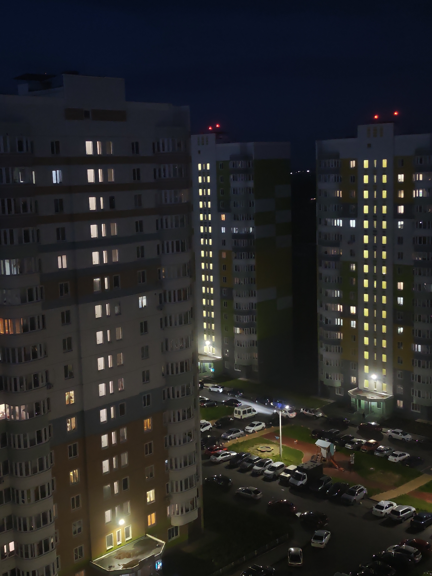 The image shows an aerial view of a residential complex at night, likely in Kursk, Russia. Three tall apartment buildings are visible, with many windows illuminated, suggesting residents are present. Red lights are flashing on top of two of the buildings, possibly for aviation safety. Below the buildings, a parking lot is filled with numerous cars, and a playground area is also visible. The overall scene depicts a quiet, urban residential setting after dark.