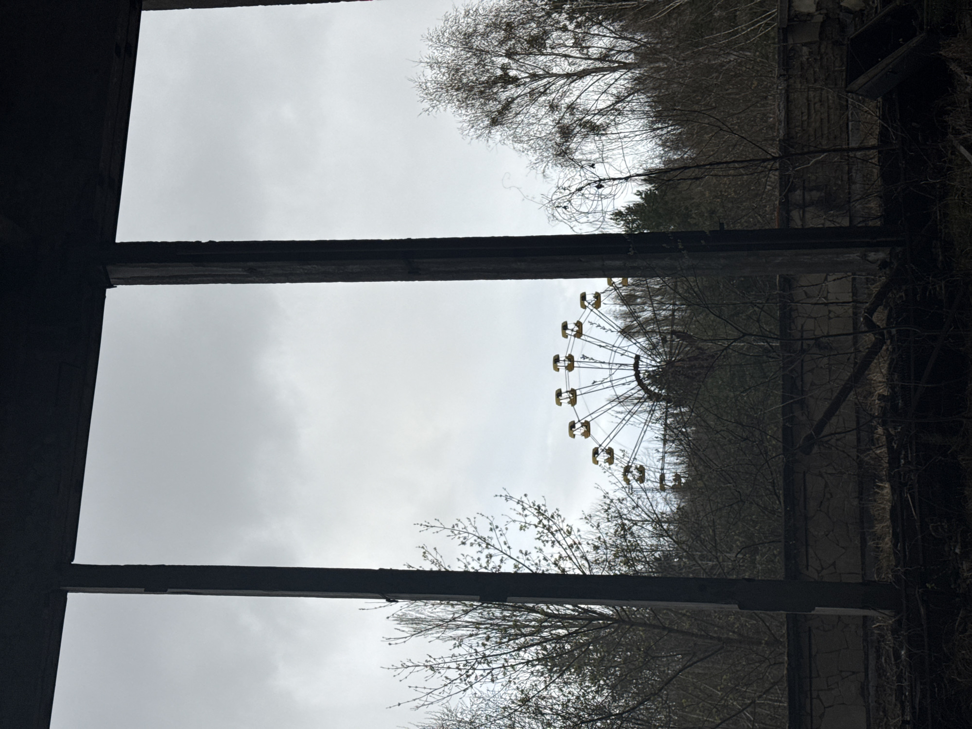 From a partially collapsed building in Prypiat, Ukraine, a Ferris wheel is visible through window frames. The sky is overcast. Bare trees surround the Ferris wheel.