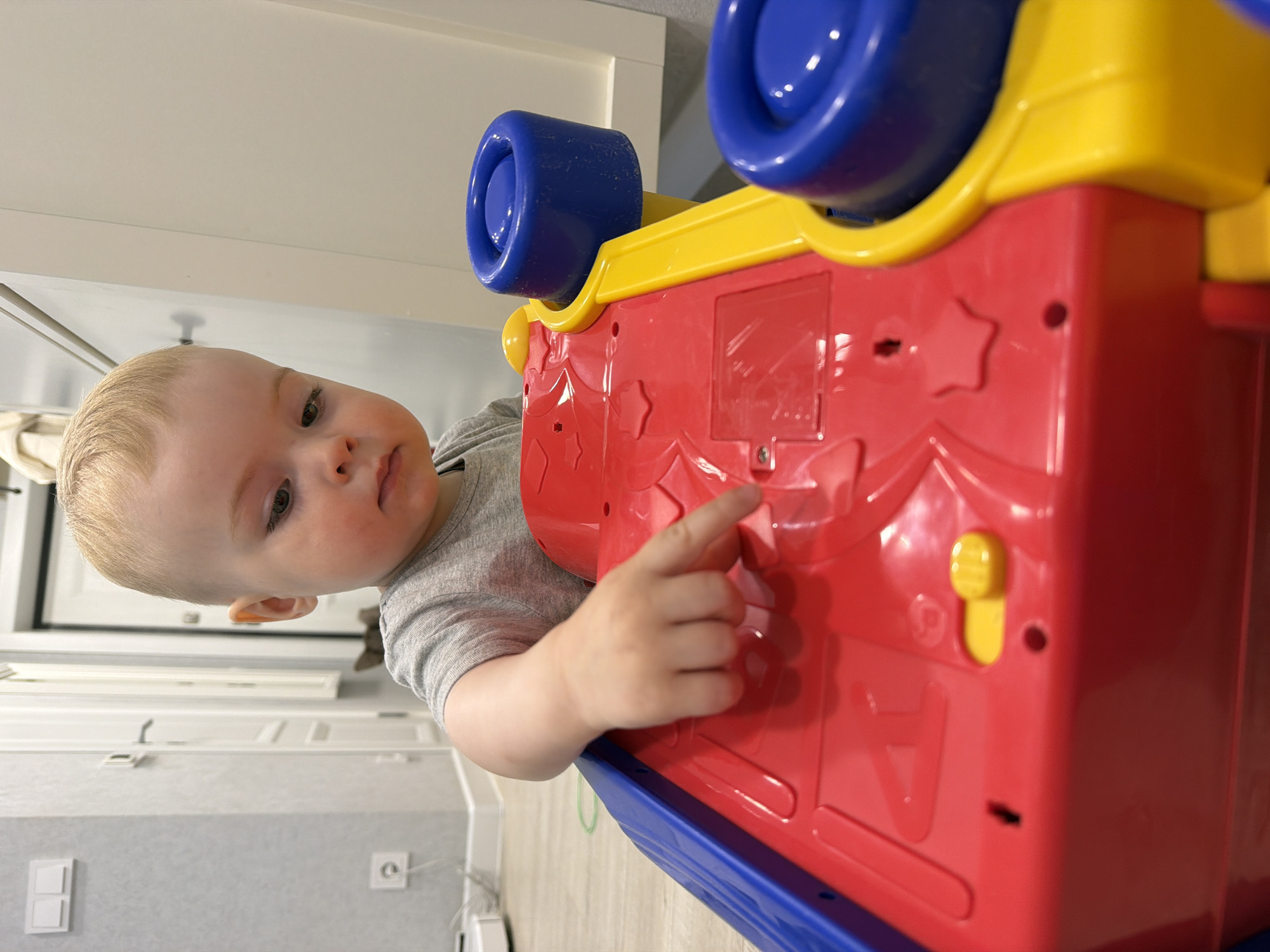 A young child, a baby or toddler with blonde hair and fair skin, is depicted in a close-up, portrait-style indoor photograph. The child, wearing a grey short-sleeved t-shirt, looks directly towards the camera with a neutral, slightly curious expression. Their right hand gently touches a brightly colored interactive toy.
The toy, prominent in the foreground, is primarily red with yellow and blue accents. It features large blue cylindrical elements on top, which might be wheels or handles, and a yellow top panel. The main red body of the toy has embossed patterns, including stars and partially visible letters "P", "A", and "B", suggesting an educational or activity toy. A small yellow knob and a circular hole are also visible on the red surface. The base of the toy is blue.
In the background, white household furniture, likely cabinets or shelves, is visible. Below these, a light grey wall features a white light switch or electrical outlet. A white door or doorframe is partially visible on the left side of the frame. The lighting is natural and even, suggesting it is daytime. The scene conveys a moment of playful interaction within a clean, modern home environment in Votkinsk, Russia.
