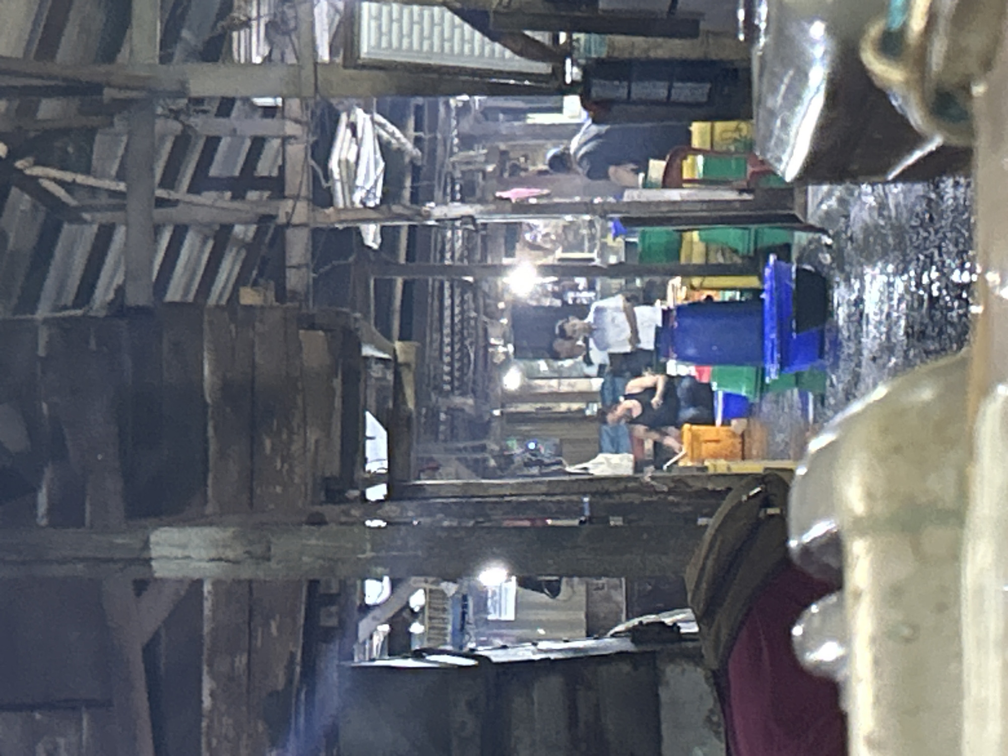 The image depicts a bustling indoor market scene, likely in North Jakarta, Indonesia, characterized by its rustic wooden structure and wet, possibly flooded, floor. In the foreground, a large, metallic object with a reflective surface dominates the lower portion of the frame. Moving further into the market, several individuals are visible. One person, wearing a black tank top, is crouched near a bright orange bucket, appearing to be engaged in an activity related to the market's trade. Behind them, a stack of green and yellow plastic crates suggests the storage or display of goods. To the left, a person in a maroon shirt and a dark backpack is seated. In the mid-ground, several other individuals stand, some in lighter colored shirts, suggesting interactions or work within the market. The environment is dimly lit, with some artificial lighting contributing to the ambiance. The overall impression is of a functional, possibly fish or produce, market with a sense of activity and a slightly weathered, utilitarian aesthetic.
