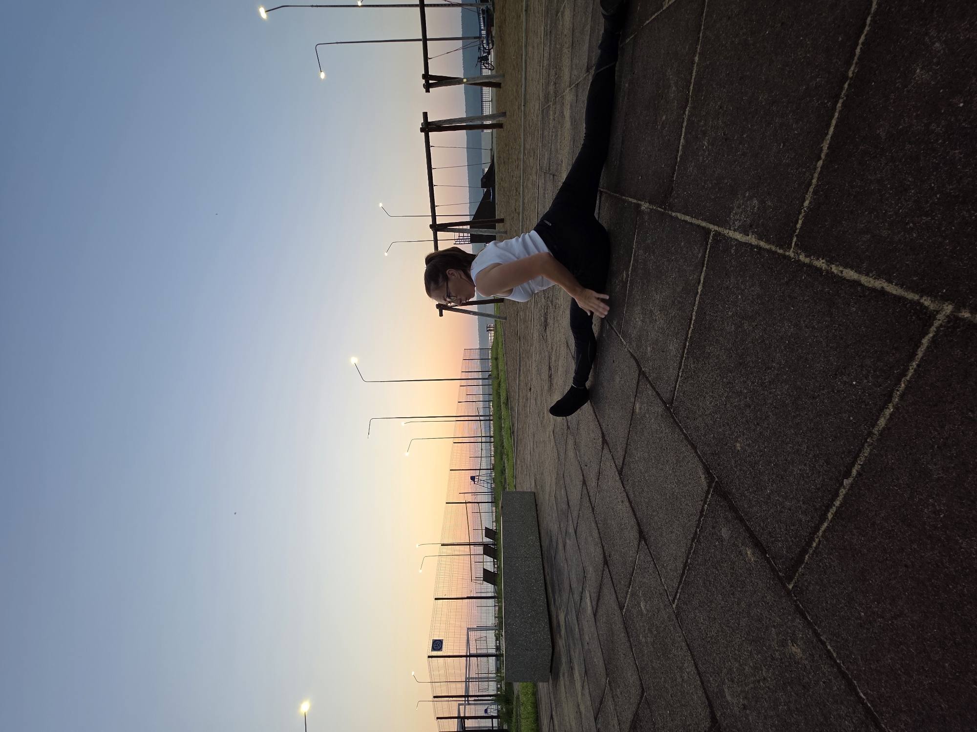 An athletic young woman with glasses and her hair in a ponytail performs a straddle split on a rubberized paved surface outdoors. She is wearing a white tank top and black leggings. Her body is positioned to the right of the frame, with her legs extended horizontally in opposite directions. Her left hand is on the ground for support, while her right arm is bent and resting on her right thigh.
The scene appears to be a public outdoor recreational area, likely a sports ground or park, possibly in the city of Chaikovsky, Russia, given the architectural style of the playground equipment and streetlights. In the background, there is a chain-link fence, and beyond that, a body of water and a tree line. Several streetlights with illuminated bulbs are visible, indicating it is either dusk or dawn. Playground equipment, including swings and a structure resembling a climbing frame, are present in the mid-ground. A long, gray concrete bench is situated to the left of the woman. The sky is a gradient of pale blue and orange, suggesting a sunrise or sunset. The overall lighting is soft, casting subtle shadows.