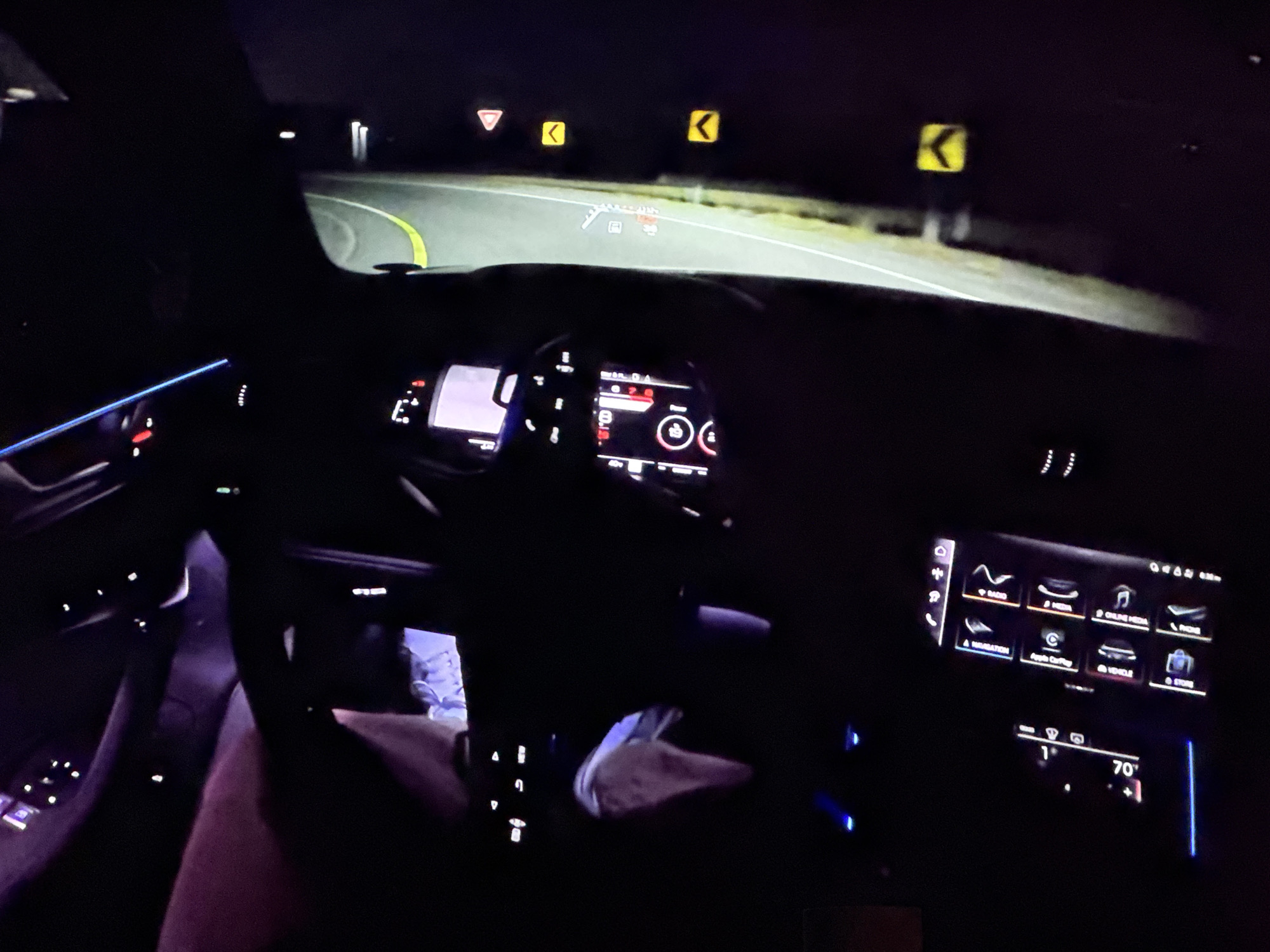 This image captures the interior of a car at night, likely in Bezden, Bulgaria, as suggested by the user prompt. The driver's perspective is shown, with the steering wheel and dashboard illuminated by ambient light and the car's internal lighting. The road ahead is visible through the windshield, curving to the right and marked with yellow lines. Several road signs are visible, including a yield sign, two chevron signs indicating curves, and a digital heads-up display projected onto the windshield showing navigational information and speed.
The dashboard features multiple screens, including a central infotainment display showing various icons for functions like radio, media, navigation, and phone. Below this, another screen displays climate control information, showing a temperature of 70 degrees. Blue LED ambient lighting is present along the dashboard and door panels. A person's legs are visible in the lower left of the frame, indicating the presence of a driver or passenger. The overall environment is dark, consistent with nighttime driving.