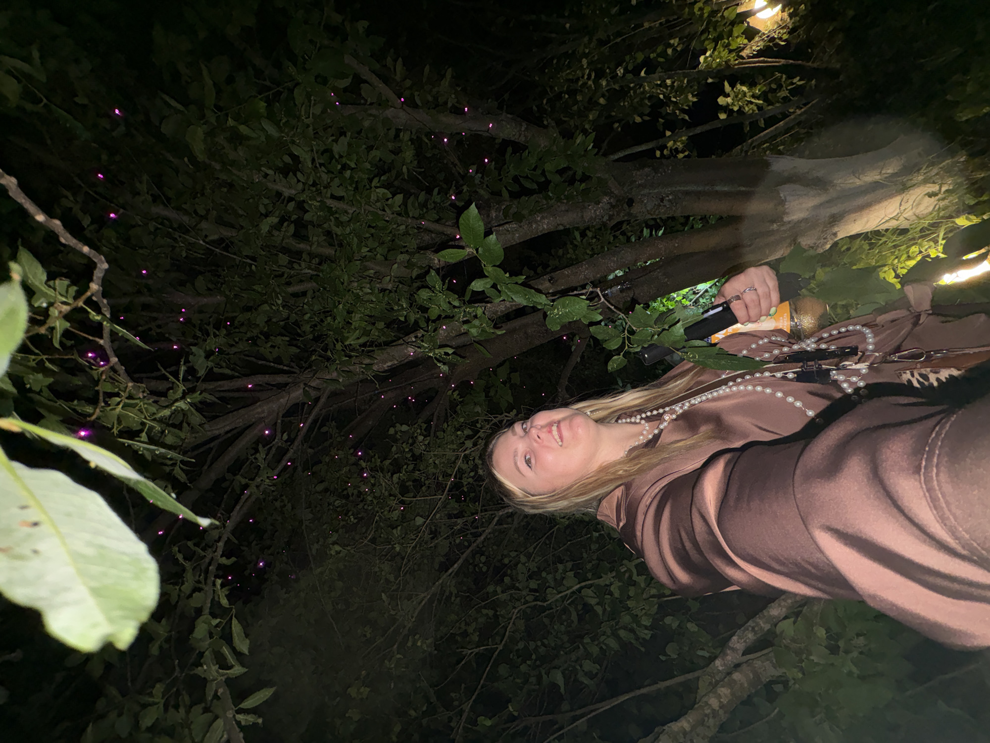 A low-angle medium shot shows a young Caucasian woman from the chest up, looking up and to her left with a slight smile. She has blonde hair and is wearing a dark brown satin shirt and a pearl necklace. In her right hand, she is holding a dark cylindrical object with an orange label. The background is a dark, wooded area with trees and foliage. Small, purple lights are scattered throughout the branches of the trees, suggesting some form of decoration or natural phenomenon. The lighting is dim, indicating it is nighttime. The location is Zavod Mikhailovskii, Russia.