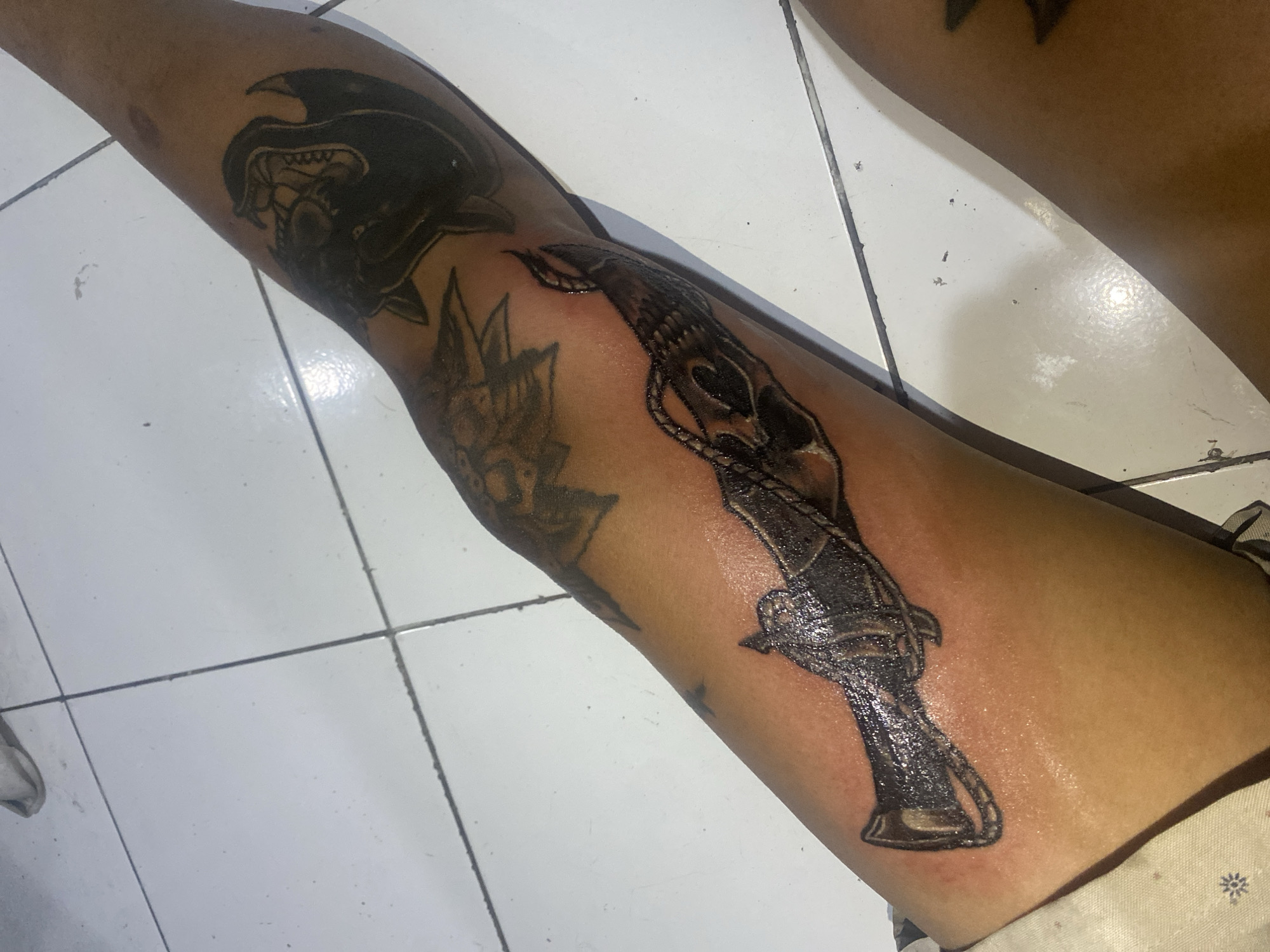 A person's lower leg, from the calf to just above the ankle, is shown resting on a white tiled floor in what is identified as Kuta, Indonesia. The leg has several tattoos. A large, dark, stylized head of a creature, possibly a panther or dragon with an open mouth and sharp teeth, is visible on the upper left side of the leg. Below this, a lighter-shaded, more faded tattoo of an abstract or floral design can be seen.
The most prominent tattoo is a large, black, intricate design resembling a ceremonial dagger or ornate weapon, positioned vertically along the shin. It features a detailed hilt with a skull-like element at the top and appears to have a rope or chain-like pattern wrapped around it. This tattoo shows signs of being very recent, with a noticeable glossy sheen, likely from ointment or fresh ink, and some redness around its edges.
The skin tone of the leg is medium to dark. The white ceramic tiles of the floor have dark grout lines and show some minor specks of dirt or debris. A small portion of light-colored, patterned fabric, possibly shorts or pants, is visible at the bottom right corner of the image. No visible text is present in the image. The scene suggests an indoor setting where tattoo work may have recently been completed or is in the healing process.