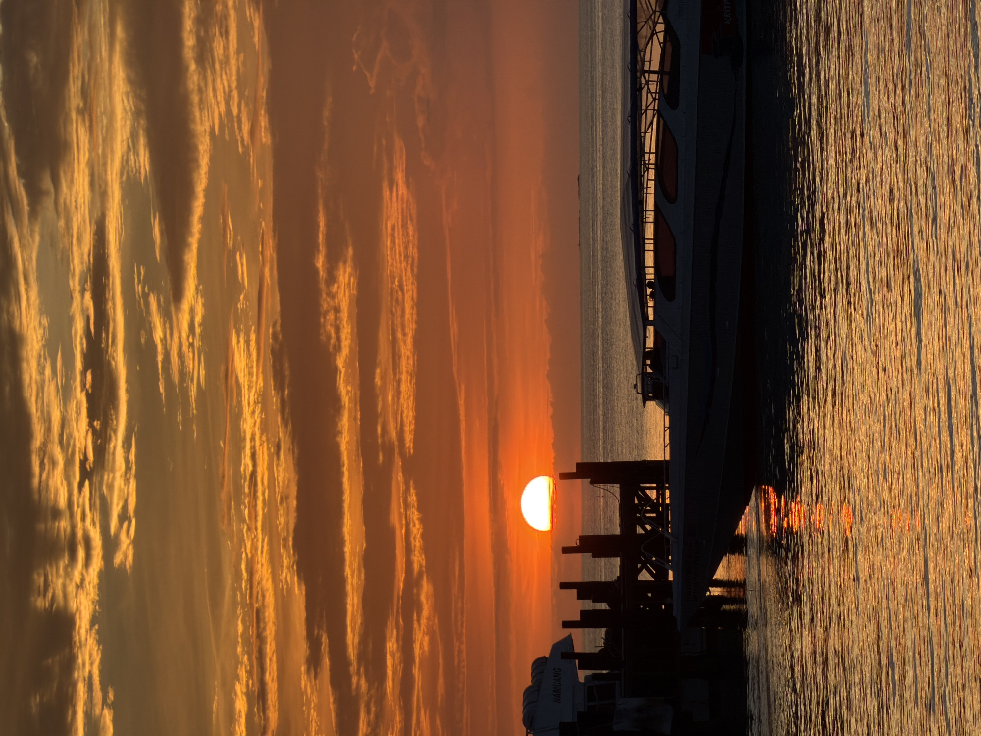 This is a vertical, medium shot of a sunset over the ocean in the Ko Samui District of Thailand. The sun, a bright orb, is setting on the horizon, casting a warm orange glow across the sky and the water. The sky is filled with colorful clouds, ranging from deep orange to light yellow. In the foreground, a boat is docked at a pier, its silhouette dark against the vibrant sky. The water reflects the colors of the sunset, creating a shimmering effect. The scene is peaceful and serene, capturing the beauty of a tropical sunset.