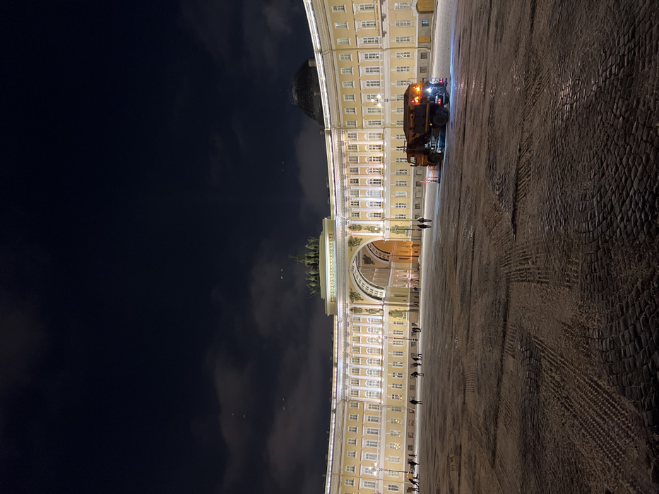 The image depicts a grand architectural scene at night, likely a large European city square. The original image is rotated 90 degrees counter-clockwise; when viewed upright, the scene reveals a long, ornate building forming a curved facade on the left side of the frame, with a vast open square on the right.
The central and most prominent architectural feature is a large, classical building, possibly a palace or a government building, illuminated with warm, golden light. It is characterized by numerous regularly spaced windows, many of which are lit, and a series of columns or pilasters along its length. A large, ornate archway with a triumphal arch style is visible towards the center-left, featuring intricate sculptural details, including a prominent statuary group on its top, which appears to depict chariots or figures on horseback. Above the archway, a dome-like structure is also visible, though partially obscured by the main building's roofline. The building's color appears to be a light yellow or cream.
The ground of the square is made of cobblestones, which appear wet and reflective, suggesting recent rain or snow/ice. Distinct tire tracks are visible across the surface, especially closer to the foreground and middle ground, indicating vehicle movement.
In the middle ground, on the right side of the square, a large utility vehicle, possibly a street-sweeper or snowplow, is parked or slowly moving. It has multiple flashing orange or amber lights on its roof and rear, typical of a maintenance vehicle. Its lights cast reflections on the wet ground.
Several small figures, presumably people, are scattered across the square, some walking, others standing. Their presence gives a sense of scale to the massive architecture.
The sky is dark night, with some soft, diffused clouds visible in the upper left portion of the image. The overall illumination comes from the building lights, the vehicle's lights, and possibly distant streetlights, creating a moody and dramatic atmosphere. No discernible text is visible in the image. The location strongly resembles Palace Square and the General Staff Building in Saint Petersburg, Russia.