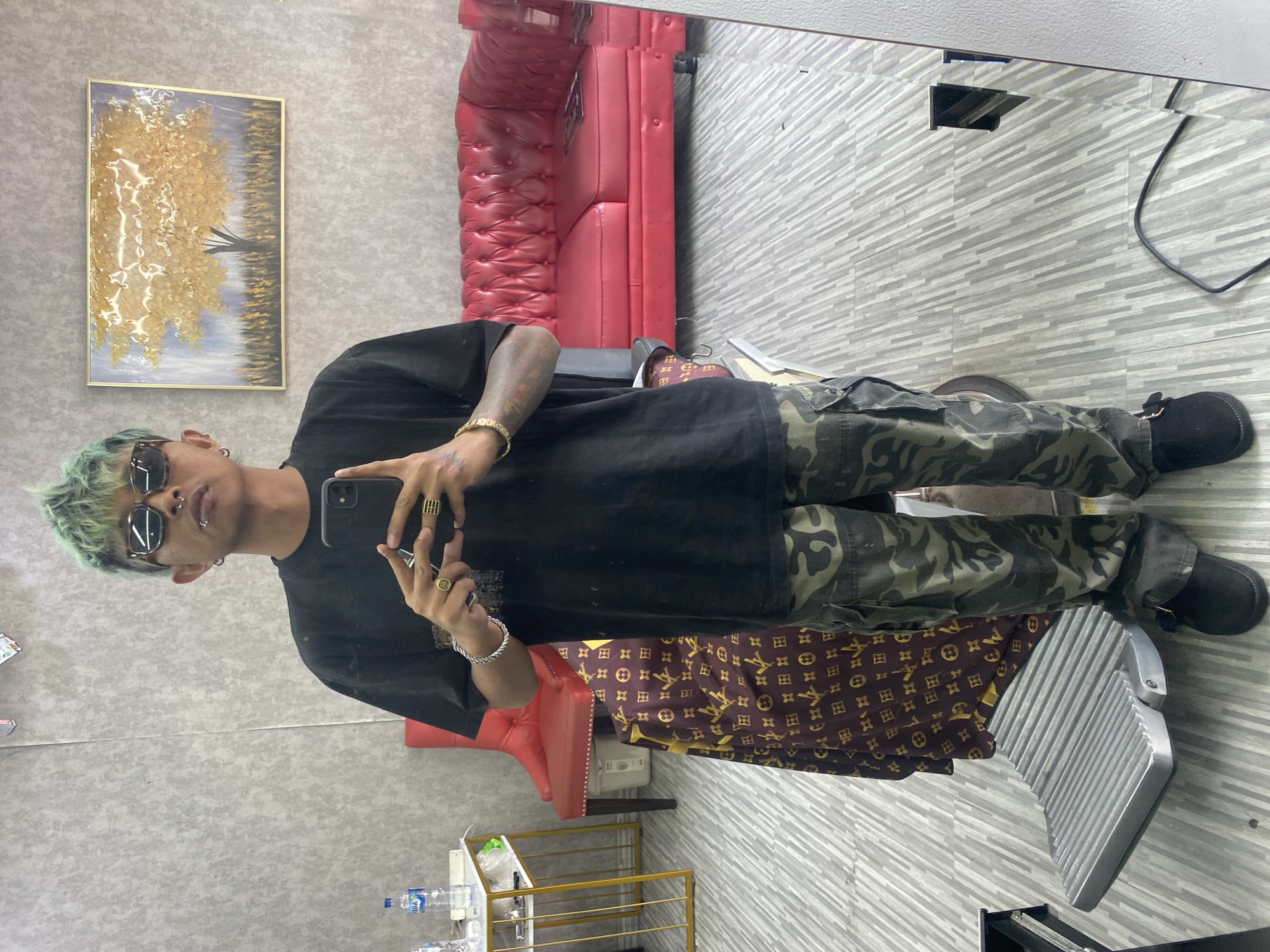 A young person, sporting light green hair, sunglasses, and facial piercings (a septum and lip ring), is captured in a mirror selfie within an indoor setting. They are wearing an oversized black t-shirt, camouflage cargo pants, and black shoes. Their fingers are adorned with multiple rings, and a gold bracelet is visible on their right wrist as they hold up a black smartphone to take the picture.
The background, reflected in the large mirror, reveals what appears to be a barbershop or salon. To the left, a vibrant red tufted leather couch or chair stands out. Above it hangs a framed painting depicting a golden, textured tree against a muted blue and grey background. The floor is covered with light grey and white patterned tiles. A brown cloth with a gold monogram pattern is draped over an unseen object near the person's legs. A silver, ribbed metal footrest is also visible. In the foreground, on a small gold-framed shelf, several bottles of "AQUA" brand water are stacked. The overall scene suggests a contemporary and stylish environment in Kuta, Indonesia.