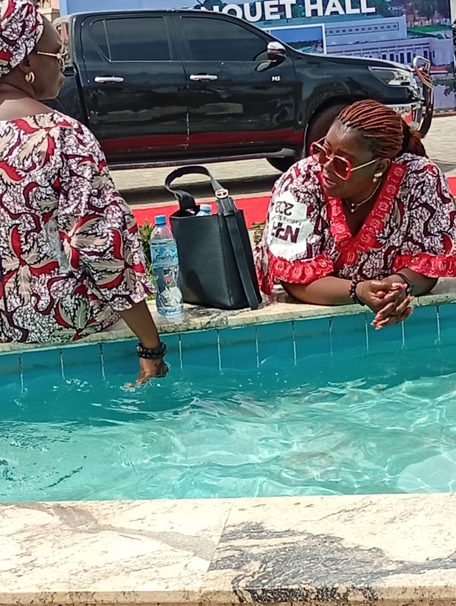 Two women are seated outdoors beside a turquoise water feature, likely a fountain or a shallow pool, in what appears to be Unguwan Sanusi, Nigeria. The scene is captured during the daytime under bright lighting.
On the left, a woman seen from behind is wearing a patterned red, white, and brown traditional garment (likely an Ankara print) and a matching headwrap. She has her right foot, adorned with dark ankle beads, partially submerged in the water, causing ripples. A black handbag and two plastic water bottles are visible on the edge of the water feature next to her.
On the right, another woman is partially visible from the chest up, leaning forward with her hands clasped. She has reddish-brown braided hair pulled back, and is wearing oversized red-rimmed sunglasses and a patterned traditional top with red trim and white accents, similar in style to the first woman's attire. A gold necklace and bracelet are also visible. On her top, there's a white patch with partial text that appears to read "202...", "Kaduna S...", and "EN", possibly indicating an event or organization related to Kaduna State.
In the background, a dark-colored pickup truck (resembling a Toyota Hilux) is parked. Behind the truck, a large white building is visible with the words "BOUQUET HALL" written on its facade, suggesting the setting is an event venue. The water feature has a light-colored, possibly granite or marble, tiled edge.