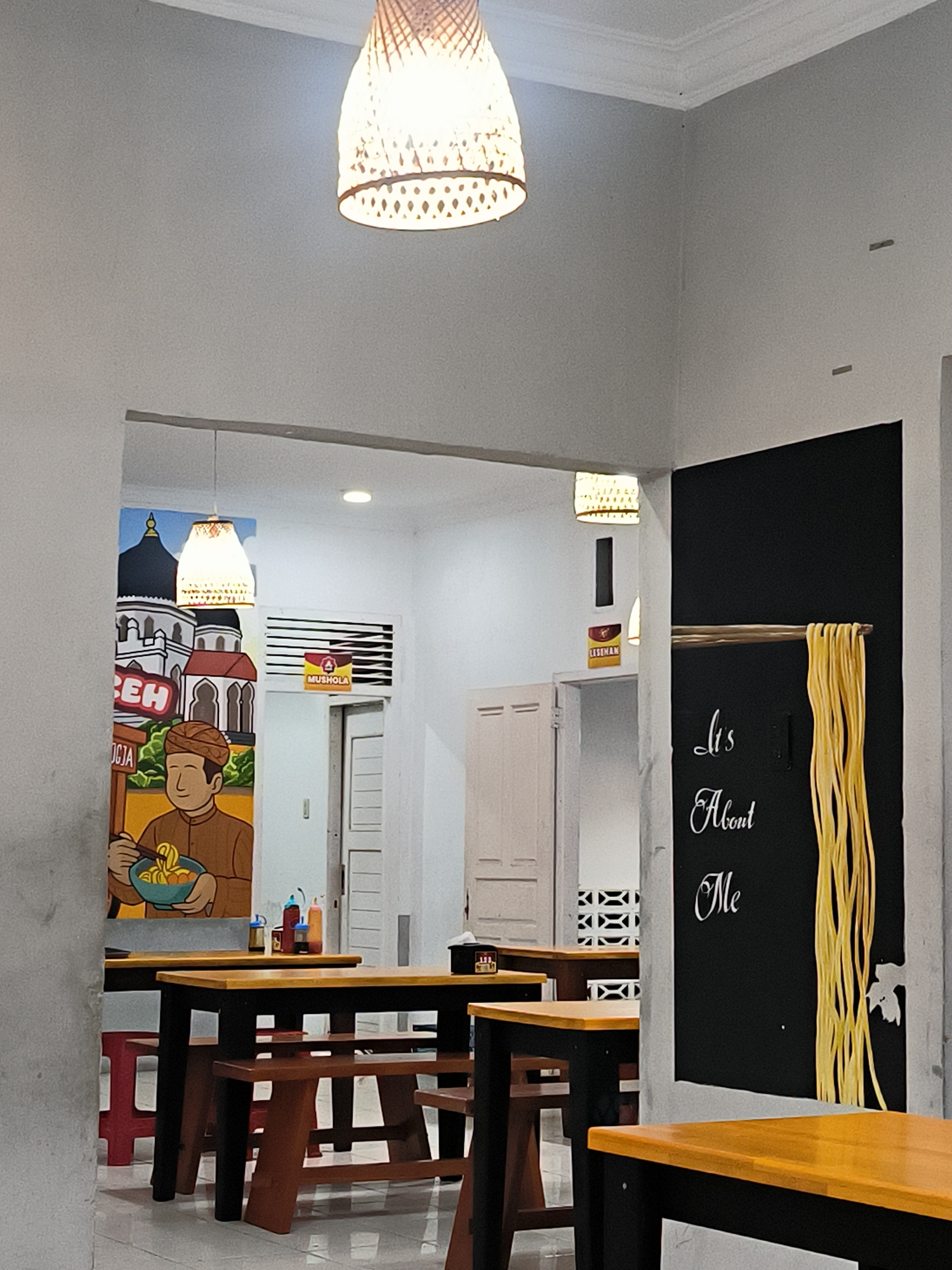 This is an interior photograph of a casual restaurant or noodle shop, likely located in Banda Aceh, Indonesia. The setting features multiple dining tables with wooden tops and dark frames, accompanied by both wooden and red plastic stools, indicating a ready-to-serve environment. No people are visible.
The walls are predominantly light gray, illuminated by several woven pendant lights and recessed lights, suggesting an indoor evening scene or a brightly lit interior. On the left side, a large, colorful mural depicts traditional Indonesian architecture, featuring mosque-like domes and minarets. The mural also includes text, with "ACEH" prominently displayed and "JOGJA" visible below. A cartoon-style man in traditional attire is shown holding a bowl of noodles with chopsticks as part of this artwork. Condiment bottles are visible on one of the tables in the foreground.
Further into the background, near a plain white door, a sign with an arrow points to a "MUSHOLA" (prayer room). Another sign reads "LESEHAN," suggesting an option for traditional floor seating, common in Indonesia. On the right side of the image, a distinctive black wall serves as an artistic feature, adorned with white cursive text that reads "It's About Me." Suspended from a wooden rod against this black wall is a three-dimensional art installation resembling long, yellow strands of noodles. The floor is tiled throughout the visible area.