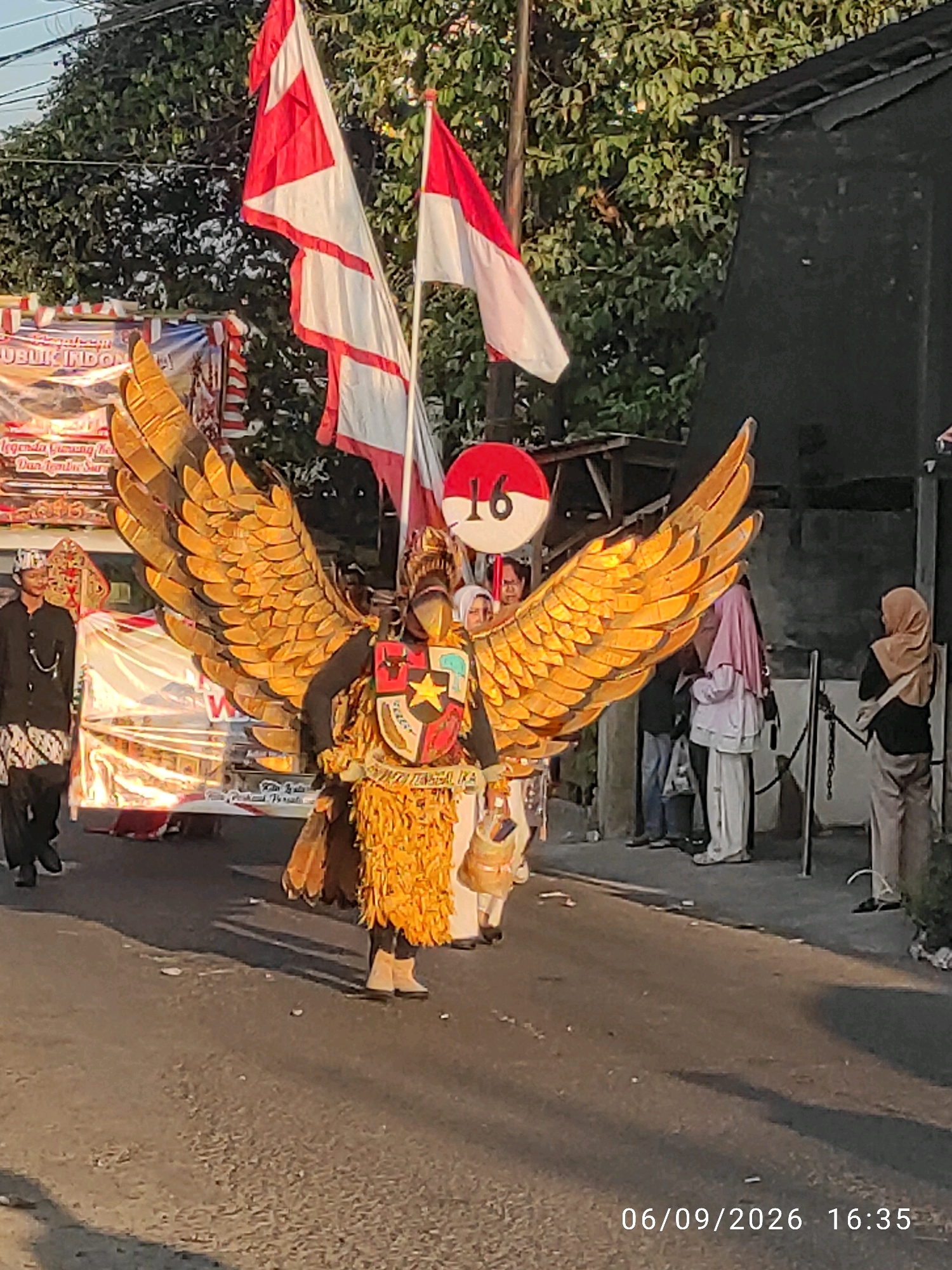 The image depicts a street scene in Wage, Indonesia, likely during a parade or cultural event. The central subject is a person in an elaborate costume resembling a golden eagle with large, outstretched wings. The costume features a decorated chest plate with symbols that appear to be nationalistic, including a red and white shield with a star. The person is wearing tan-colored boots and a textured, layered costume.
To the left of the eagle-costumed individual, two men in traditional attire are visible. One man wears a black jacket and pants with a batik-patterned sarong, and a patterned head covering. He is looking towards the right. Another person, partially obscured, is also on the left, possibly part of the parade or a spectator.
Behind the eagle costume, two Indonesian flags, red and white, are prominently displayed. One flag is larger and unfurled, while the other is smaller and partially furled. A red circular sign with the number "16" in white is positioned near the flags.
In the background, other people are seen on the right side of the street, some wearing headscarves. A dark, covered structure, possibly a building or stall, is present. A black metal fence runs along the right side of the street. The street itself is paved, with some debris on the surface. The scene is illuminated by natural light, suggesting it is daytime, with shadows cast on the road. A banner with text in Indonesian is visible on the left, partially obscured by the eagle's wing, with "PUBLIK INDONESIA" being a readable part. The date "06/09/2026 16:35" is superimposed on the lower right corner of the image.