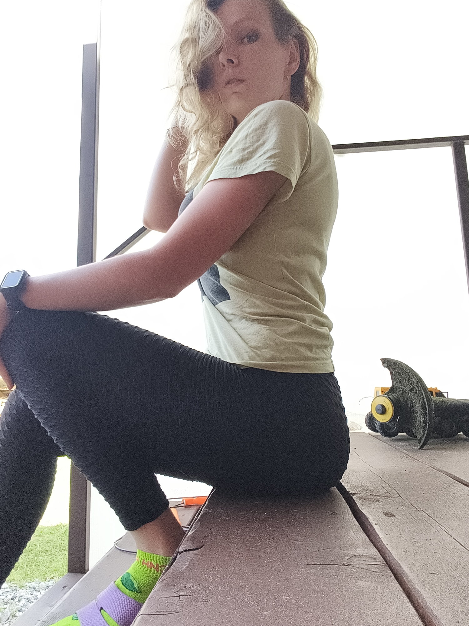 A young woman with blonde, curly hair is sitting on a wooden deck. She is wearing a light green t-shirt and black leggings. She has a smartwatch on her left wrist. Her socks are brightly colored, with green and purple accents. In the background, there is a blurry yellow toy tractor with a large cutting attachment. The scene appears to be outdoors, possibly on a porch or deck. There is a railing behind the woman. The sky is bright and overcast. The location is identified as Русский Вожой, Russia.