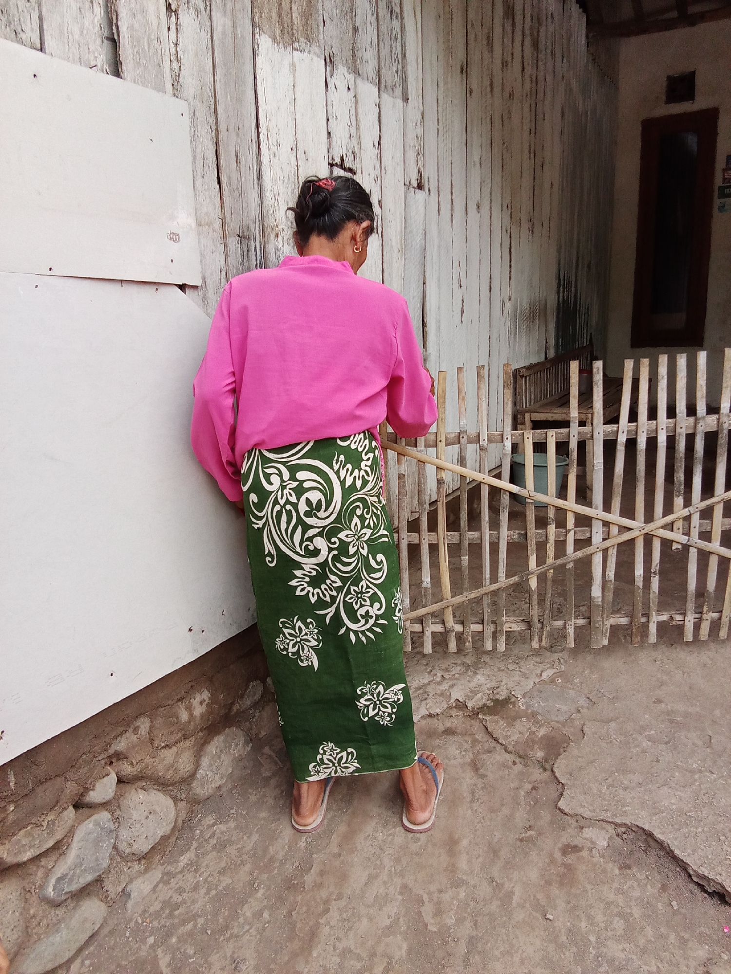 The image captures a woman from behind, standing outdoors in what appears to be a rural or less developed area. She is wearing a bright pink long-sleeved shirt and a long, dark green skirt with a white floral pattern. Her hair is pulled back in a bun, and she is wearing small hoop earrings. She has on blue and white flip-flops.
The background consists of a weathered, light-colored wooden wall on the left, possibly part of a building. To the right of the woman is a rustic fence made of vertical bamboo poles with horizontal bamboo supports. Beyond the fence, a dark wooden structure, possibly a bench or part of a building, is visible, along with a green bucket. The ground is a mix of concrete and dirt, with some small stones and cracked surfaces. The overall setting suggests a simple, outdoor environment, characteristic of a village or residential area in places like Trebungan, Indonesia. There are no other people visible in the image. The lighting appears to be natural daylight, with soft shadows.
