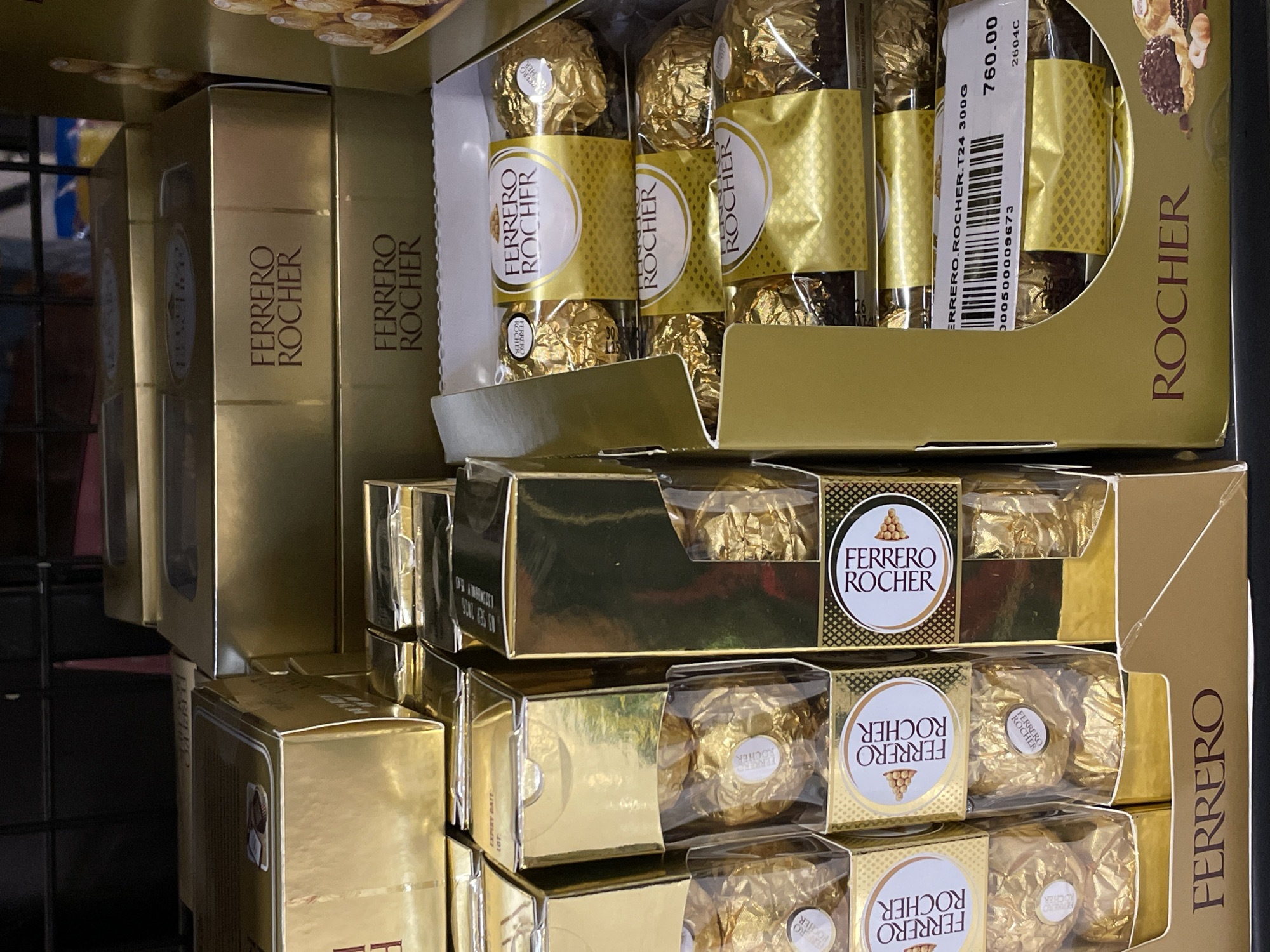 The image shows a close-up, eye-level view of a display shelf filled with Ferrero Rocher chocolates in a retail store. The chocolates are primarily presented in their signature gold foil packaging and gold-colored boxes. Multiple boxes of various sizes and configurations are stacked and arranged, some opened to reveal the individual chocolates inside. The lighting is bright, creating reflections on the shiny surfaces of the packaging. The background is slightly out of focus, showing the dark shelving structure of the display. There are no people clearly visible in the image. The scene suggests a product display for sale, likely in a supermarket or confectionery store. The presence of a price tag indicating "760.00" and product information such as "FERRERO.ROCHER.T24 300G" confirms the retail context. Without additional visual cues like store signage or specific architectural details, pinpointing the exact location within Davao City, Philippines, from this image alone is not possible.