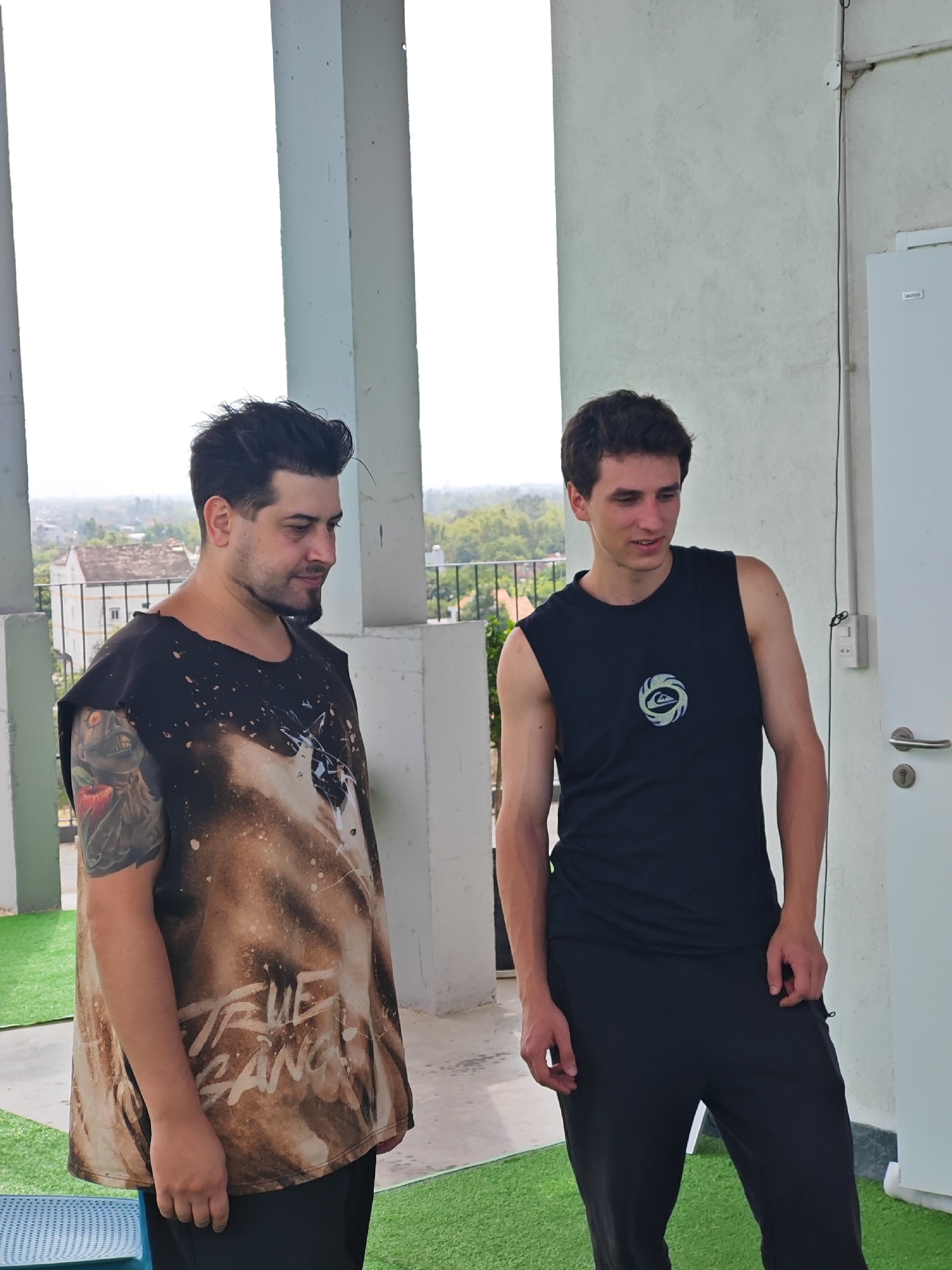 Two young men stand on a balcony, overlooking a residential area with trees and buildings. The man on the left has dark hair, a beard, and is wearing a dark t-shirt with a distressed graphic design and tattoos on his left arm. The man on the right has light brown hair and is wearing a black sleeveless shirt with a circular logo and black pants. They are both looking away from the camera. The balcony has white pillars, and a white wall is visible on the right side of the frame with a door and electrical fixtures. Green artificial turf covers the floor of the balcony. The scene is likely in Đà Nẵng, Vietnam, based on the urban and tropical landscape visible in the background.