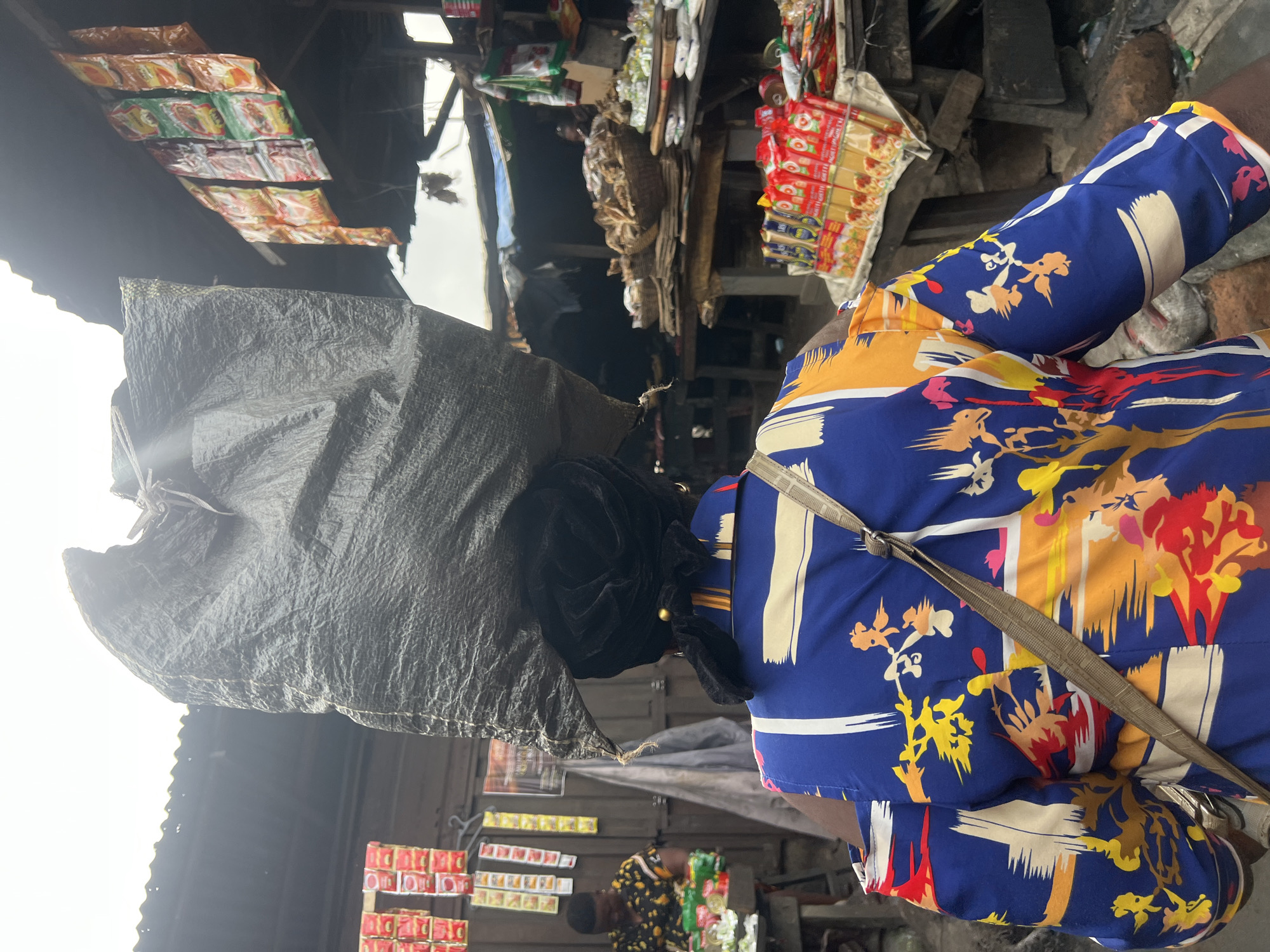 The image depicts a woman in Nkpor, Nigeria, carrying a large, dark gray plastic bag on her head. She is walking away from the camera and is wearing a vibrantly colored blue shirt with a bold, abstract pattern of yellow, red, and orange. A beige strap from a bag or satchel is visible across her right shoulder. She has a black head covering and a black ribbon tied around her bun.
The scene appears to be an outdoor market. To the left, there are shelves displaying small red and yellow packets, possibly food items or spices. Another woman is partially visible in the background, wearing a patterned shirt and attending to goods on a stall. To the right, market stalls are laden with various goods, including baskets of dried fish and stacks of packaged food items such as spaghetti. The structures overhead appear to be made of corrugated metal roofing, suggesting a covered or partially covered market area. The overall environment is bustling and indicative of a busy marketplace.