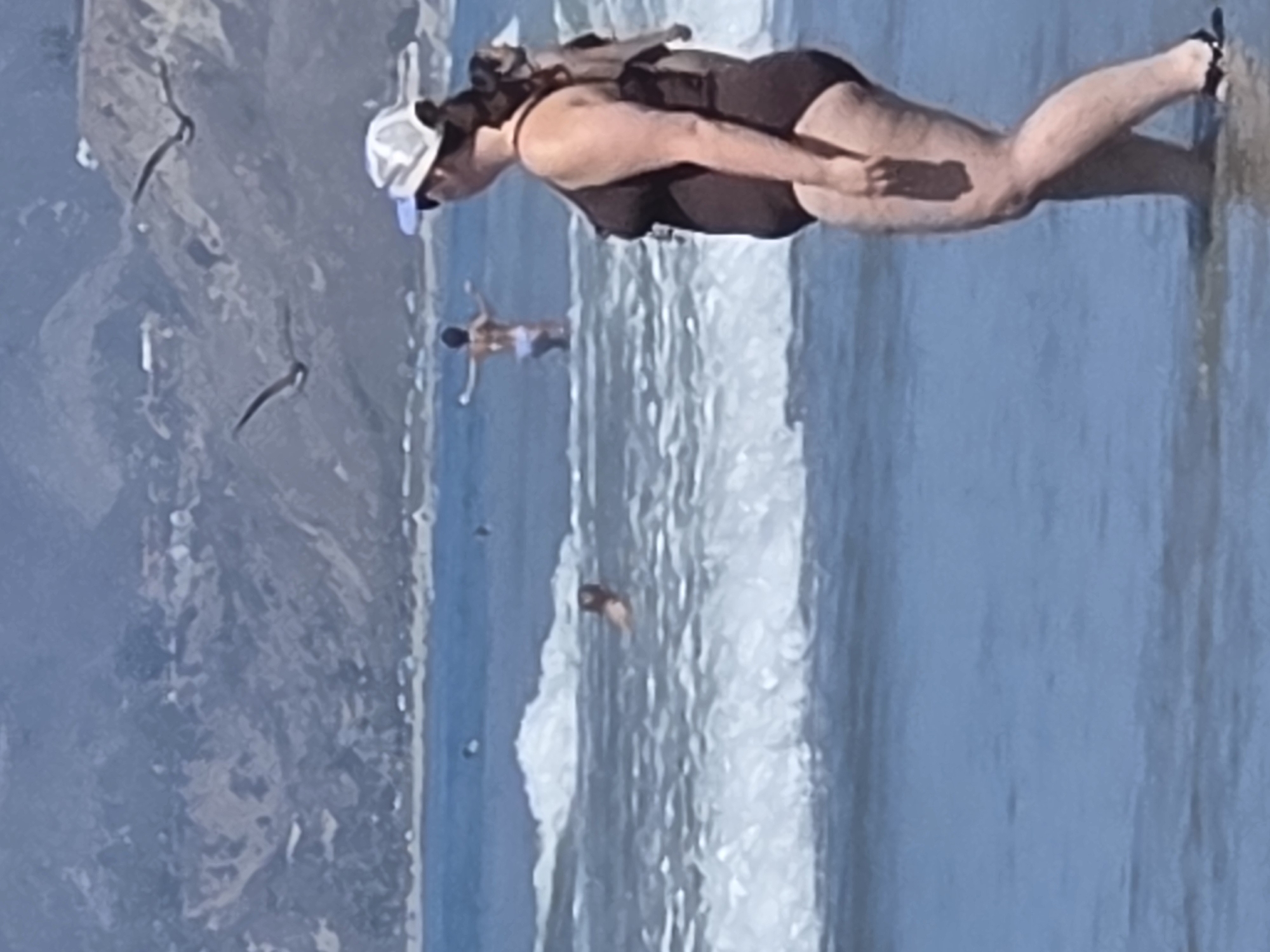 The image depicts a crowded beach scene in Santa Monica, California. In the foreground, a woman wearing a dark swimsuit and a white cap stands in shallow water, facing away from the camera. Her body is angled to the right, and she appears to be looking towards the ocean. In the middle ground, several people are visible in the water. One person is swimming, with their arms outstretched. Further out in the water, another individual is standing. Two seagulls are flying in the sky, with a backdrop of coastal hills and vegetation. The ocean has moderate waves, and the sky is clear and bright, suggesting a sunny day. The overall impression is one of a lively and popular beach destination.