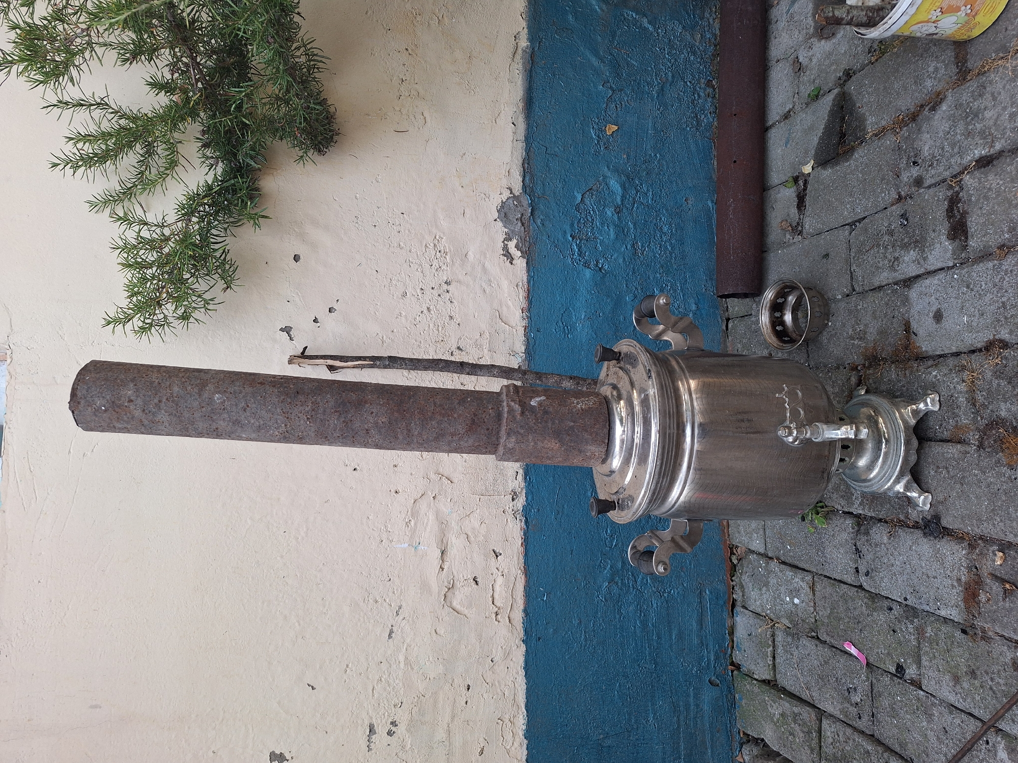 In Nabran, Azerbaijan, a weathered, silver samovar stands on a cobblestone patio, its tall, rusted chimney pipe extending upwards. The samovar has ornate handles and a spigot. A small, metal strainer sits on the ground next to it. Behind the samovar, a wall is painted in two horizontal bands: the upper section is a faded cream, textured and peeling in places, while the lower section is a vibrant, textured blue. A sprig of green rosemary grows from the cream section of the wall, its branches reaching towards the right. To the right of the samovar, a rusty metal pipe lies horizontally on the ground. In the lower right corner, a yellow bucket with a floral pattern contains a bundle of sticks.