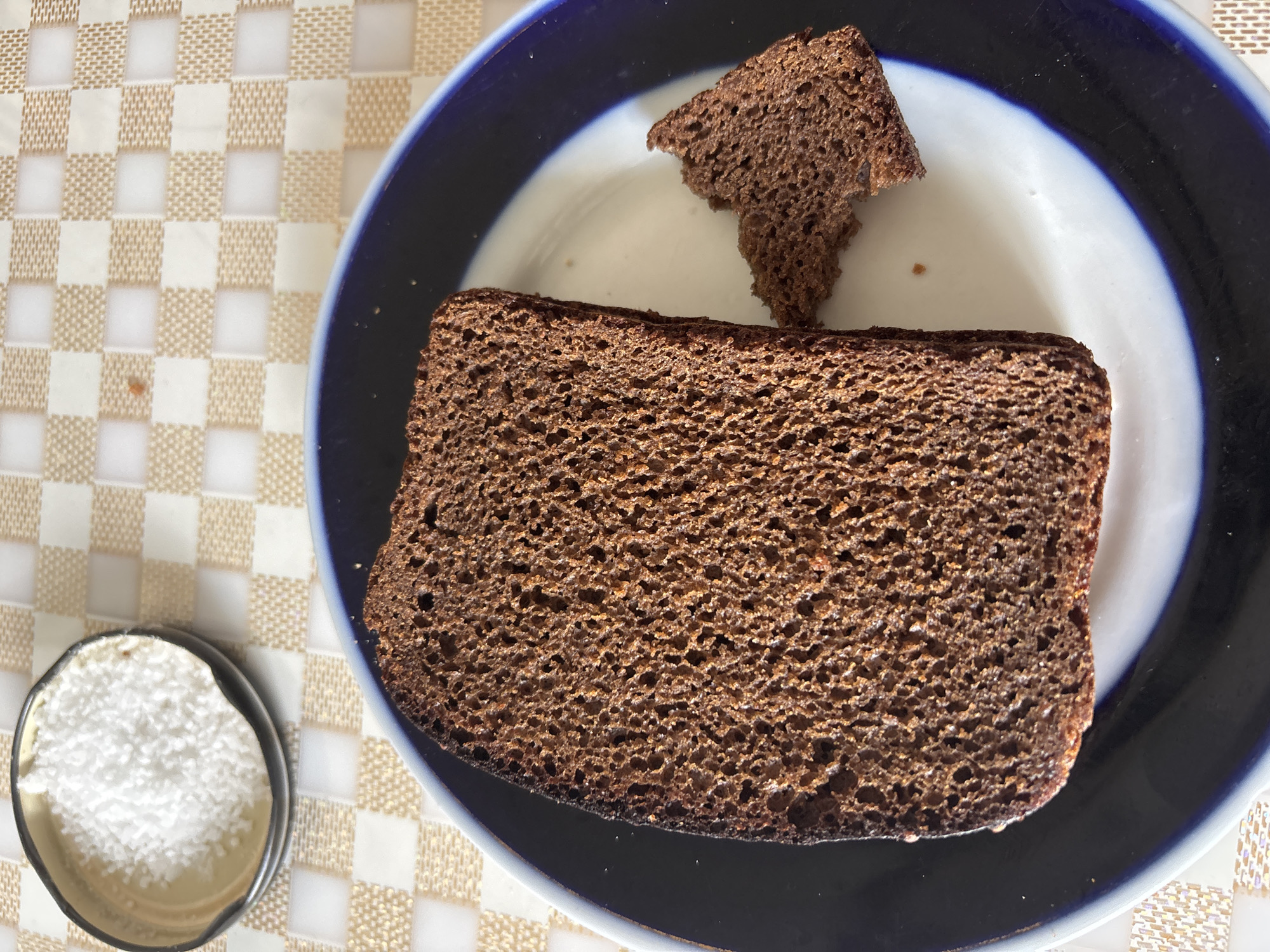 The image shows a close-up, top-down view of two slices of dark rye bread on a plate. The larger slice is rectangular and positioned horizontally, while a smaller, irregularly shaped piece is to its top. The bread is dark brown with a visible crumb texture. The plate has a dark blue rim and a white inner surface. To the left of the plate, there is a small, shallow bowl filled with a white granular substance, possibly salt or sugar. The items are placed on a surface with a checkered pattern of white and light brown squares. The lighting suggests it is daytime, with natural light illuminating the scene. The setting appears to be a dining area. There is no visible text in the image.
