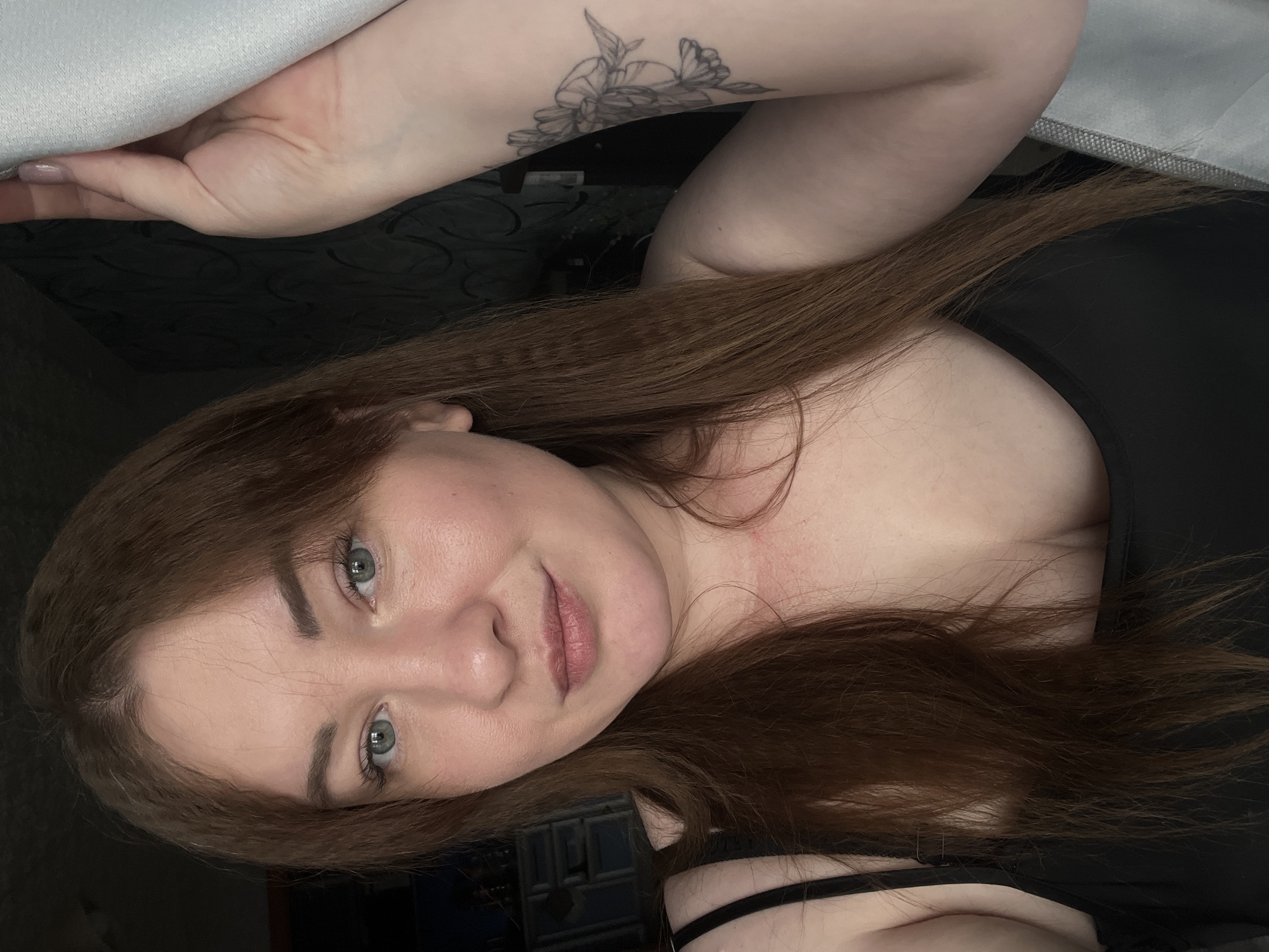 A young woman with long, reddish-brown hair and blue eyes is seen at close range. She is fair-skinned and has a floral tattoo on her left forearm. She is wearing a black top with thin straps. The background is dark and includes what appears to be a black patterned surface and a gray fabric or surface near the top of the frame. There is no evidence indicating the city of Chaikovsky, Russia, in this image.