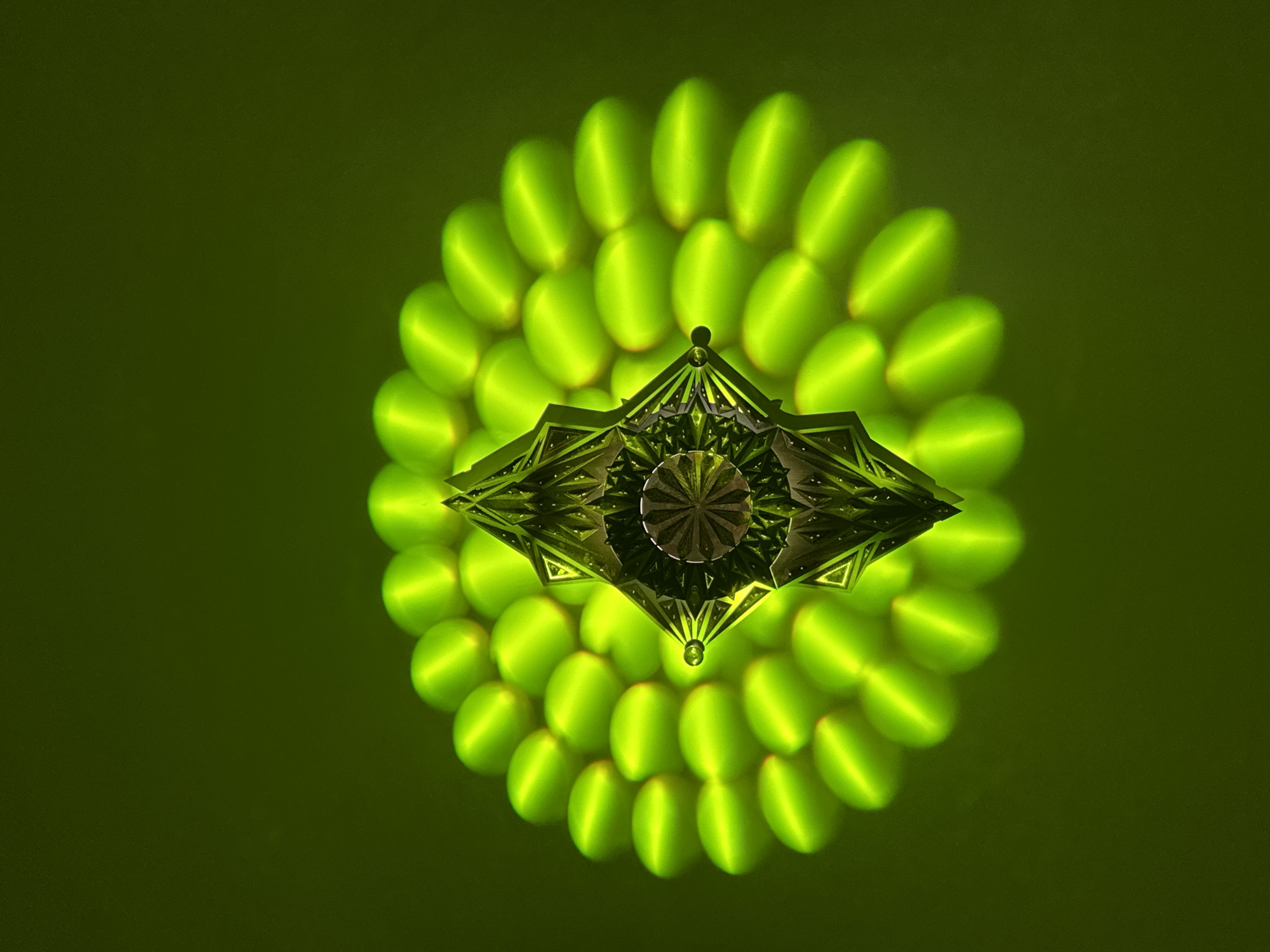 An abstract, low-angle, interior shot features a geometric light fixture composed of mirrored, diamond-shaped panels surrounding a central, circular element with a radial design. The fixture is illuminated with a bright, green light that reflects and refracts, creating a repeating, oval pattern of light around it. The background is a solid, matte green. There are no people visible in the frame.
