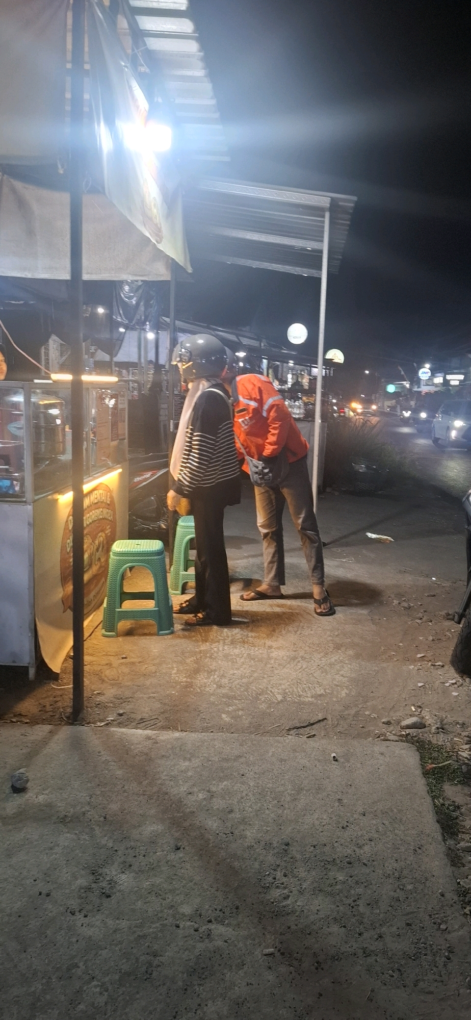 This is a nighttime photograph taken in Pabelan, Indonesia, capturing two individuals standing outside a food stall. The stall is illuminated by a bright light, casting shadows on the concrete ground. One person, wearing a helmet and a striped shirt, stands near a green plastic stool. The other person is wearing an orange jacket and is leaning towards the first individual. The background shows a street with some traffic and other illuminated stalls or buildings. The overall scene depicts a casual street-side encounter in a commercial area.