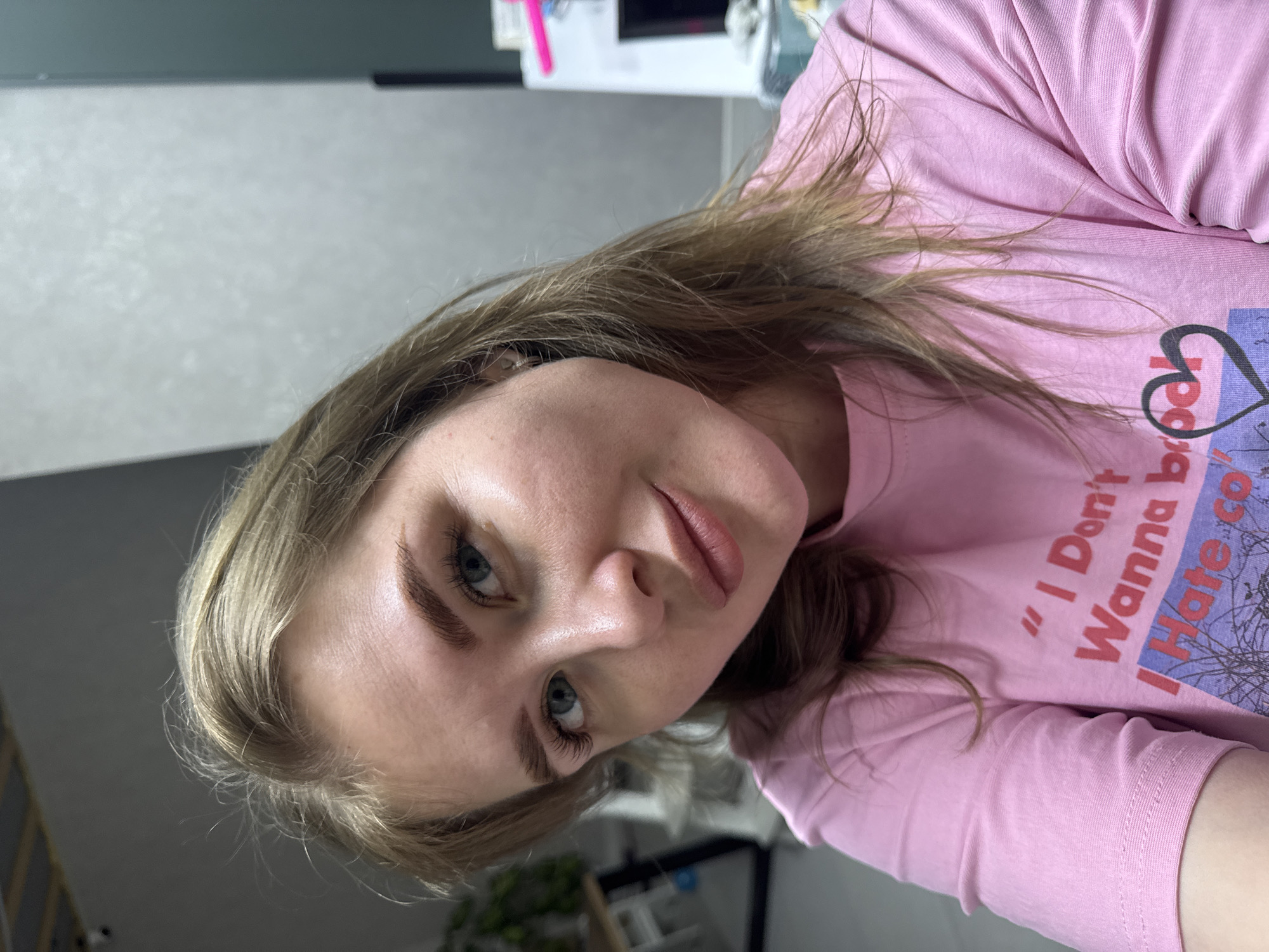 A woman with fair skin and light brown hair is looking directly at the camera. She has blue eyes and well-defined eyebrows. She is wearing a pink t-shirt with a print that reads "I Don't Wanna Hate" in red lettering. The background includes a gray wall and a glimpse of furniture and items that might be in an indoor setting, possibly in Gornyi (Dvurechenskoe S/P), Russia. The overall setting appears to be indoors with casual, everyday elements.