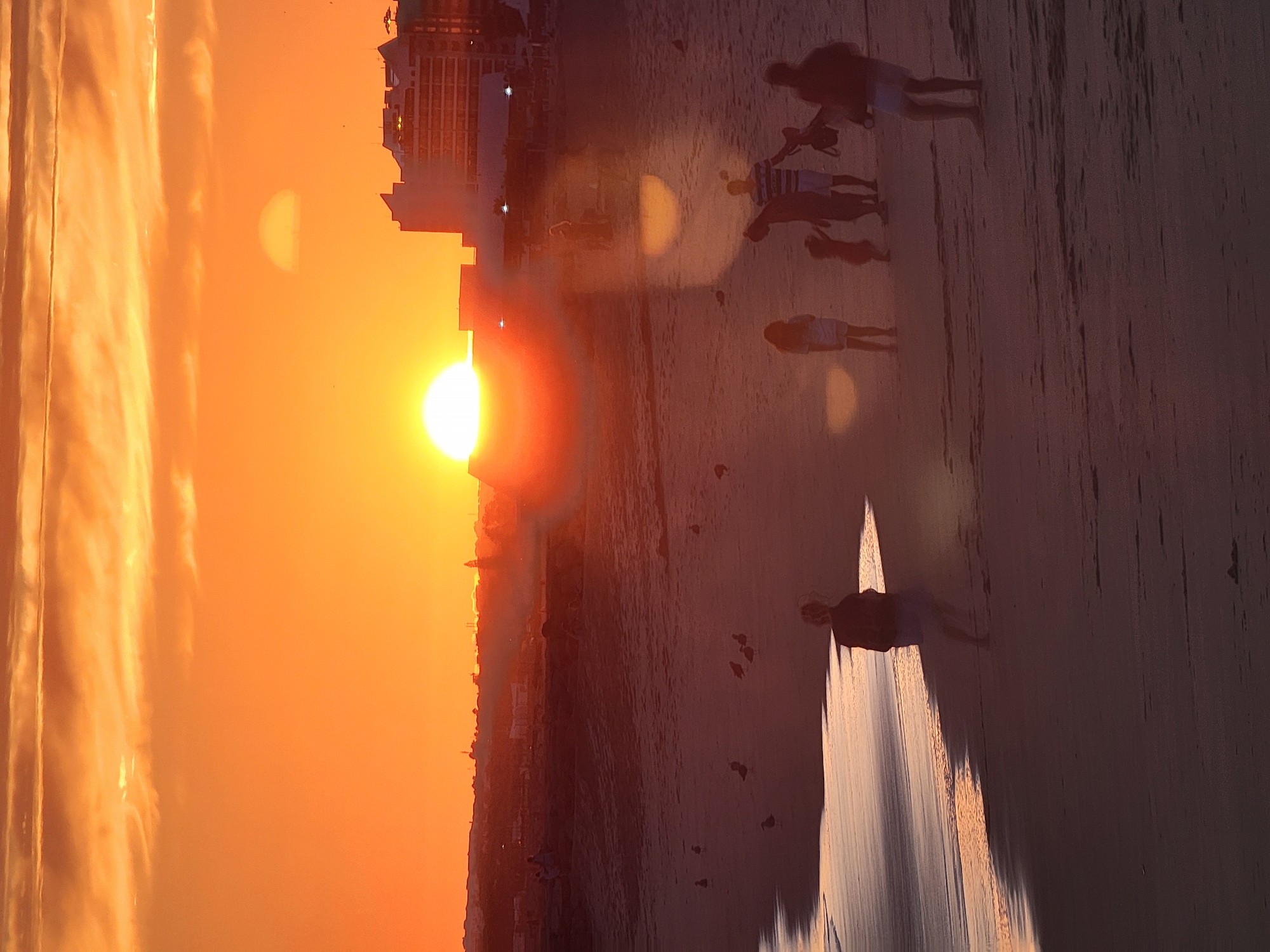 The image captures a sunset over Quarteira, Portugal. A vibrant orange sky dominates the scene, with a large, bright sun positioned low on the horizon. Clouds are scattered across the sky, reflecting the warm tones of the sunset.
In the foreground, a sandy beach stretches out, with gentle waves lapping at the shore. Several figures are present on the beach, silhouetted against the bright sky. A person walks near the water's edge, facing away from the camera. Further inland, a group of people, including adults and children, are gathered. Some appear to be walking, while others stand together. A baby stroller is visible within the group.
In the background, the cityscape of Quarteira is visible, with buildings and structures silhouetted against the sunset. The architecture suggests a coastal resort town. The overall atmosphere is peaceful and serene, characteristic of a beach sunset.