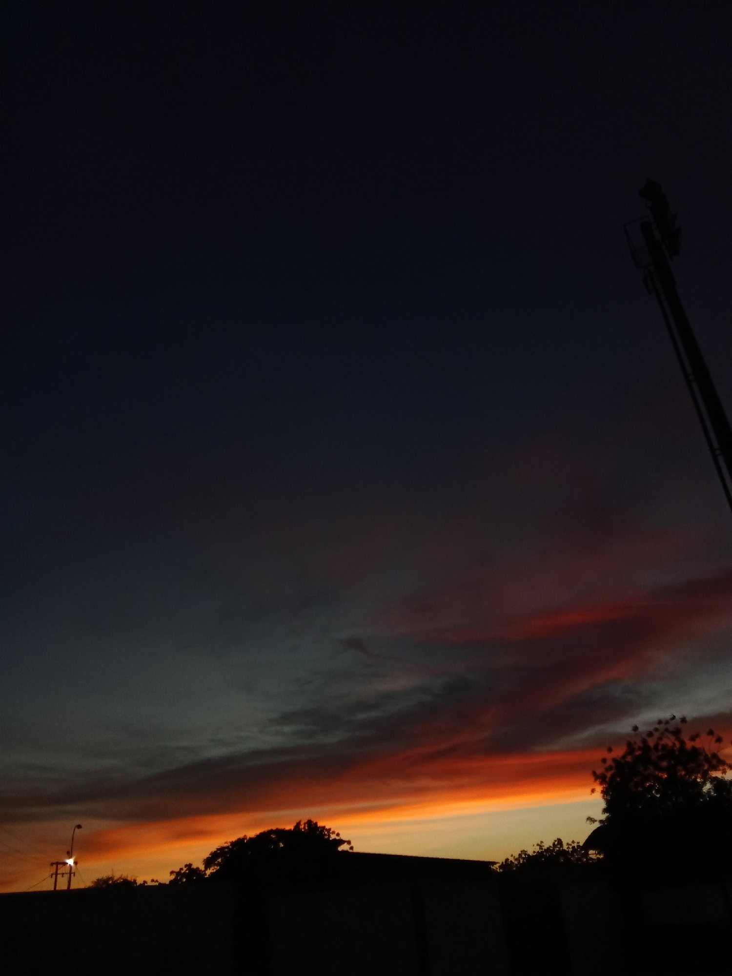 The image depicts a dark, low-angle view of a sunset sky over Juan Griego, Venezuela. Silhouetted trees and a building are in the foreground. In the lower left, there is a lit street lamp and an electrical distribution pole. A communications tower is partially visible in the upper right of the frame. The sky transitions from a dark blue-black at the top to a mix of grays and orange-red hues near the horizon, where bands of clouds are visible. The lighting suggests dusk or early evening.