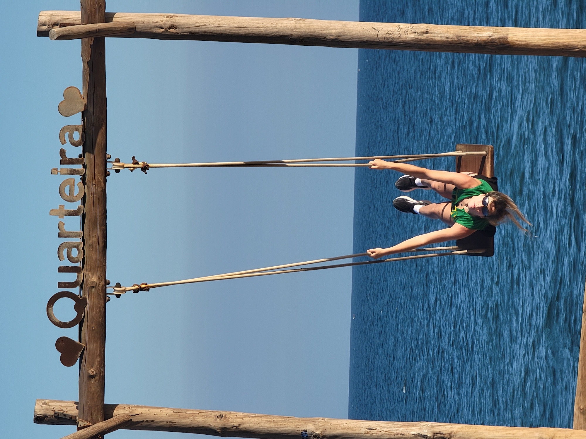 A young woman with blonde hair and sunglasses is upside down on a swing set at the edge of the water. She is wearing a green top and black shorts, with black and white sneakers. The swing set is made of wood and has a sign that says "Quarteira" with heart shapes on either side. The ocean is blue and calm, with a clear sky above. The scene appears to be in Quarteira, Portugal, a coastal town known for its beaches and tourism.